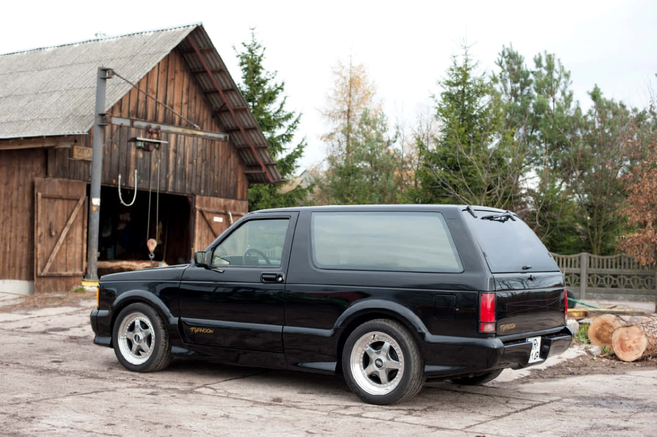 Gmc Typhoon 桌布 壁紙 — 一台時尚的黑色薩博9000 Turbo停泊在碎石庭院中，旁邊是質樸的木製穀倉，背景有樹木。