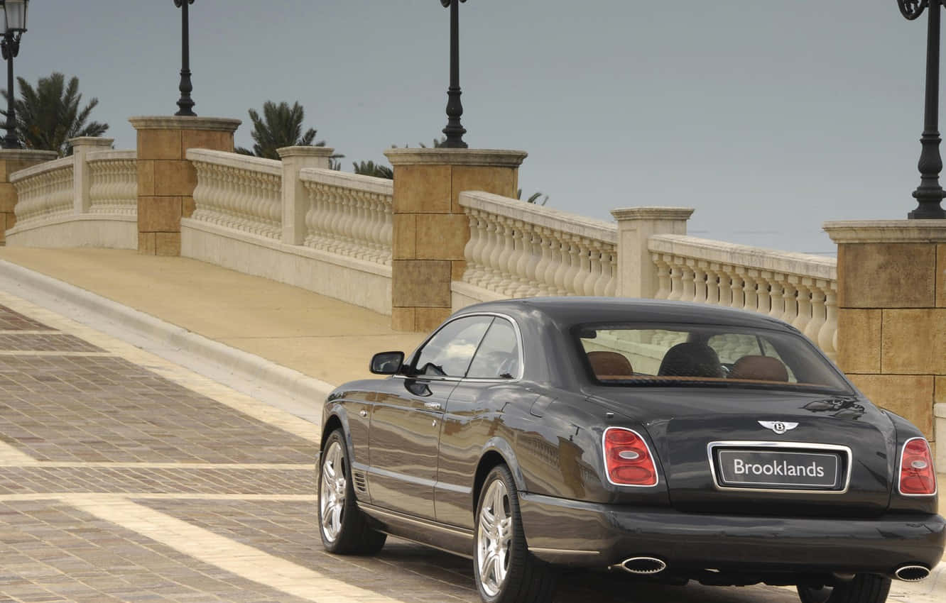 Bentley Brooklands 桌布 壁紙 — 一台時尚的黑色賓利 Brooklands 雙門轎跑車在陽光明媚的石橋上行駛，背景有華麗的欄杆和棕櫚樹。