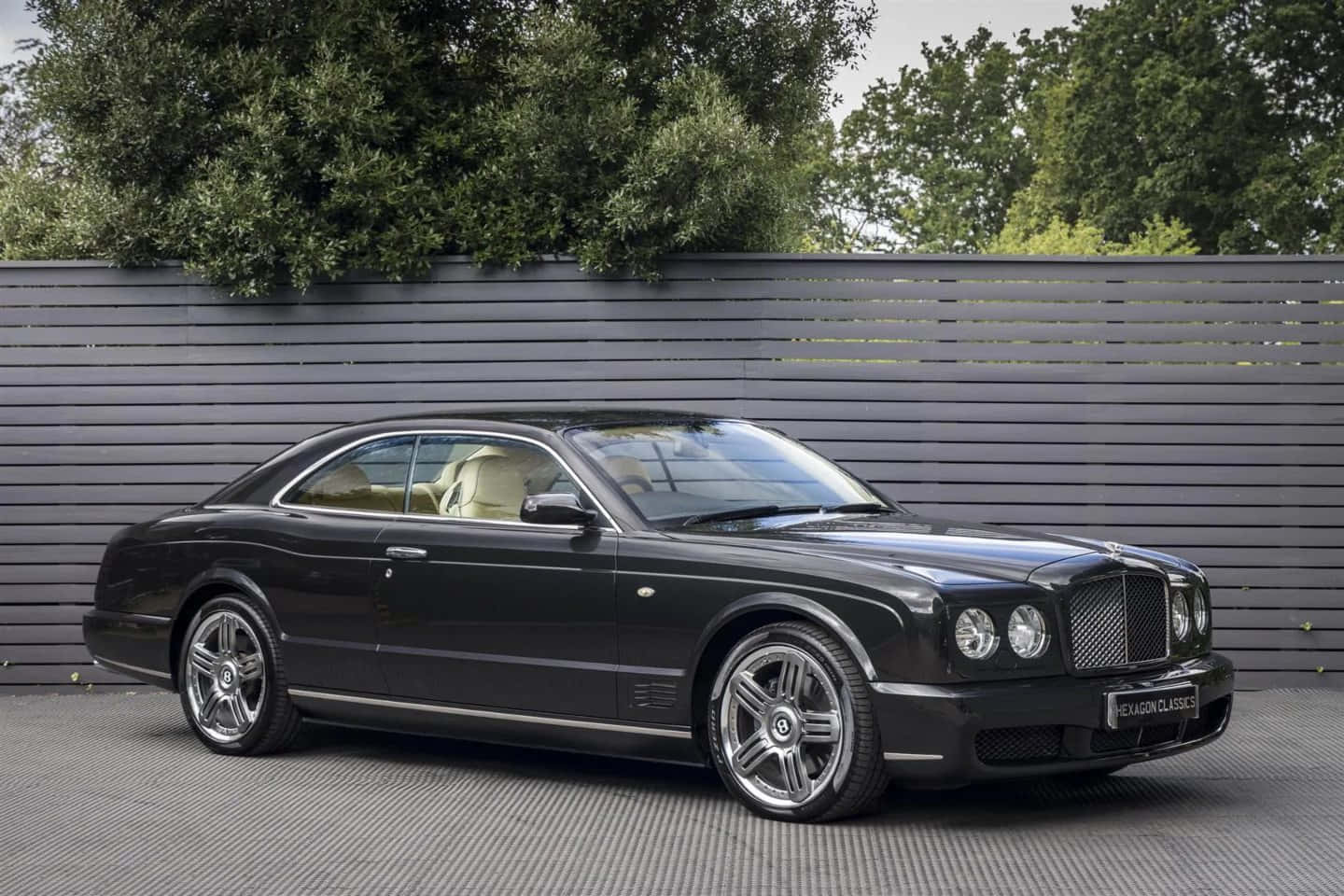 Bentley Brooklands 桌布 壁紙 — 一台時尚的黑色賓利 Continental GT 雙門跑車停在灰色網格地板上，後面有深灰色板條牆和綠色樹木。