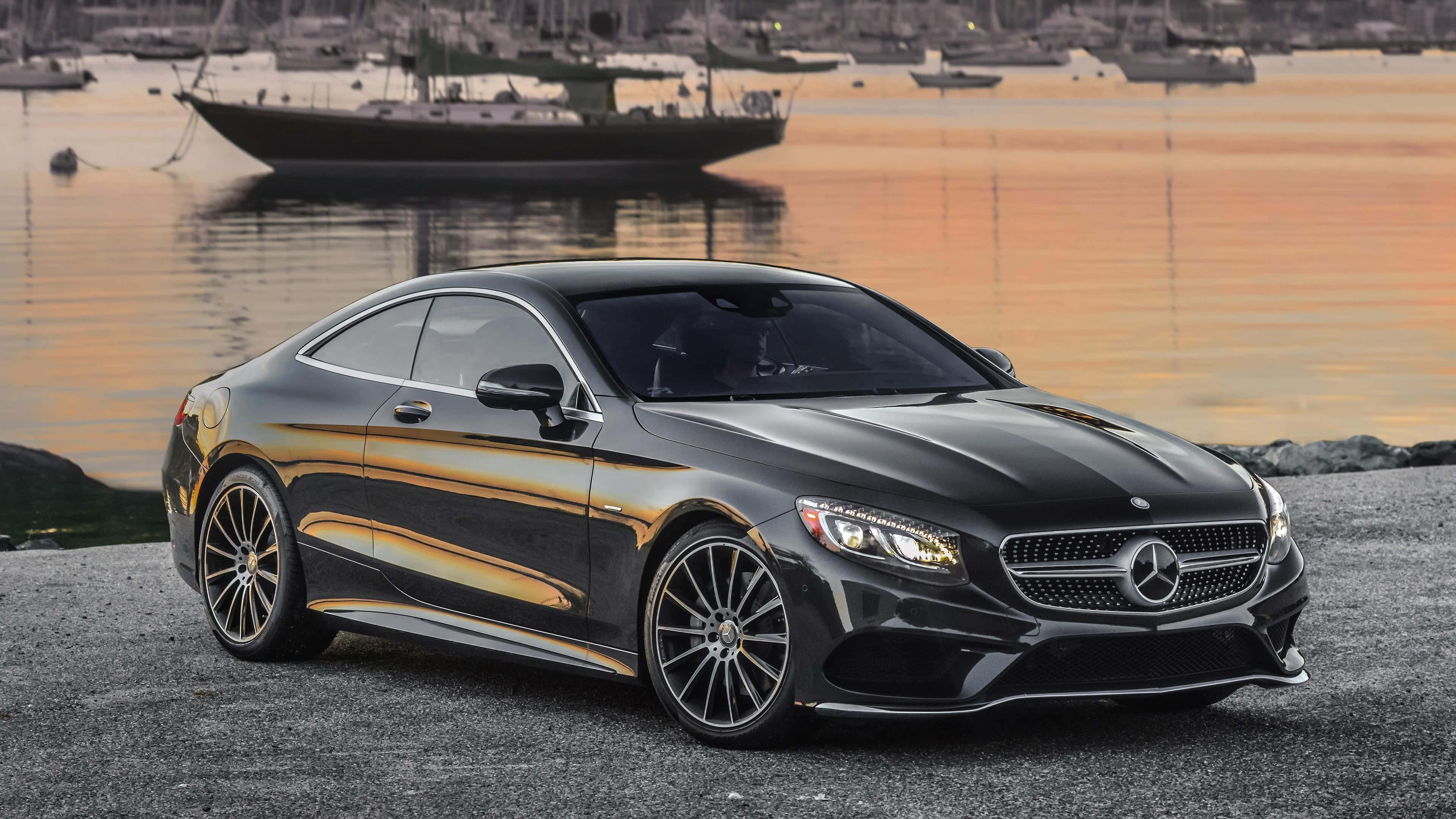 Mercedes 桌布 壁紙 — 一台時尚的黑色賓士跑車在礫石岸邊閃耀，背後金色夕陽的光線在水面上反射。