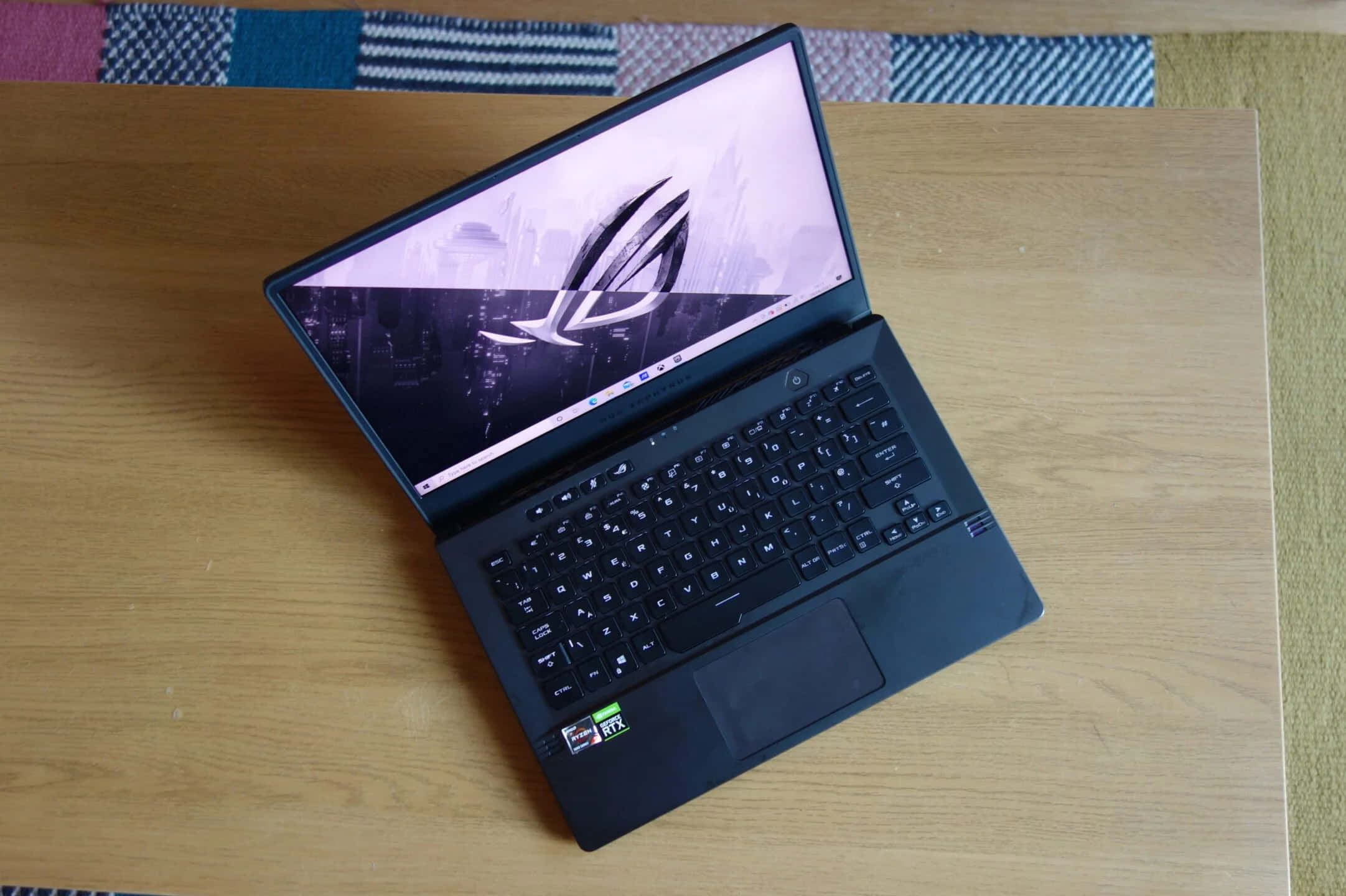 ASUS 桌布 壁紙 — 一台時尚的黑色遊戲筆記型電腦打開在淺色木桌上，顯示帶有發光ROG標誌的城市景觀桌布。