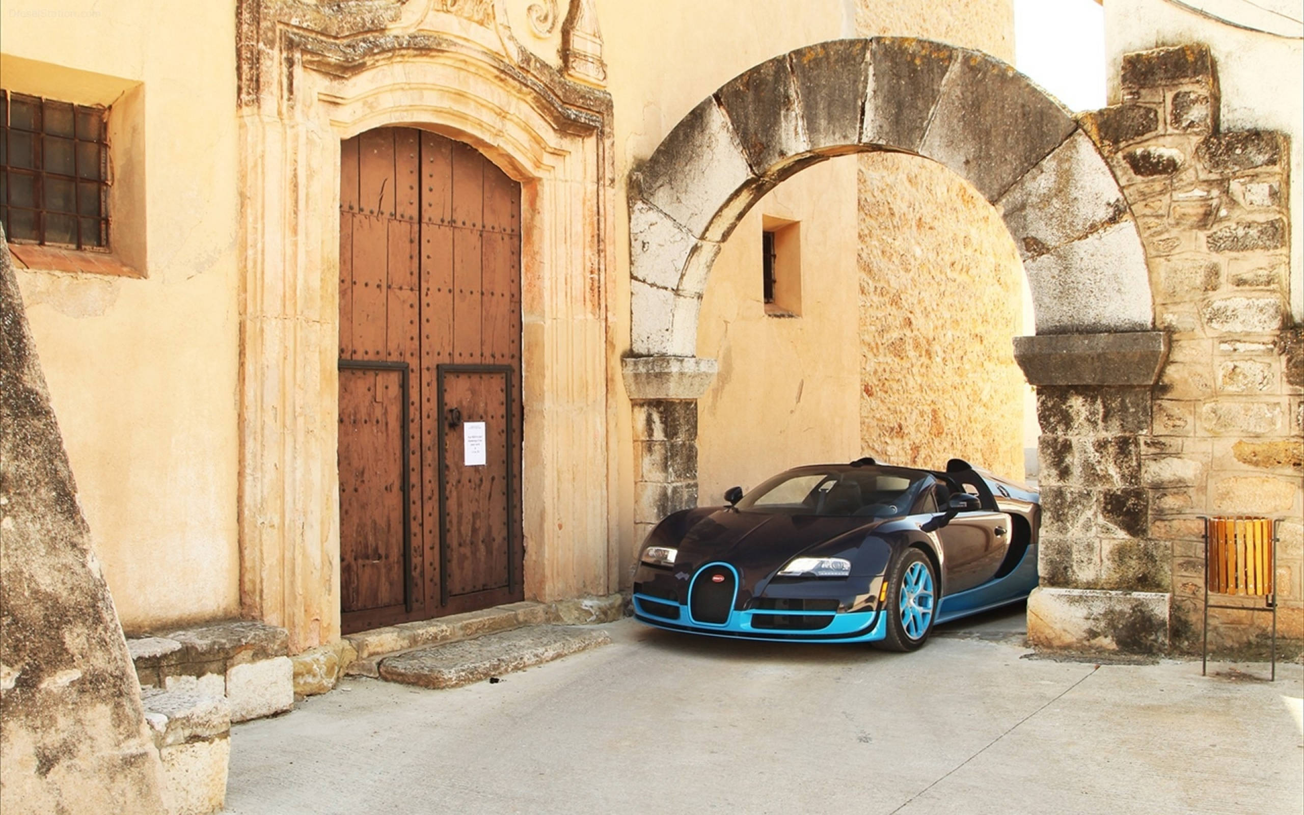 bugatti iPhone 桌布 壁紙 — 一台時尚的黑藍色Bugatti Veyron停放在風化的石拱門下，旁邊有一扇舊木門，位於陽光普照的庭院中。