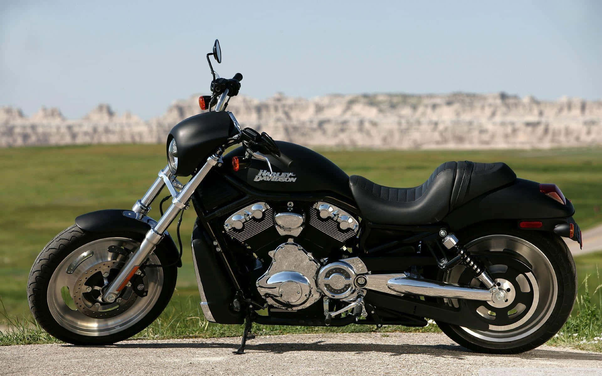 機車 桌布 壁紙 — 一台時尚黑色Harley-Davidson機車停在鋪砌道路上，背景是草地平原和遠處的岩石地貌，天空晴朗。