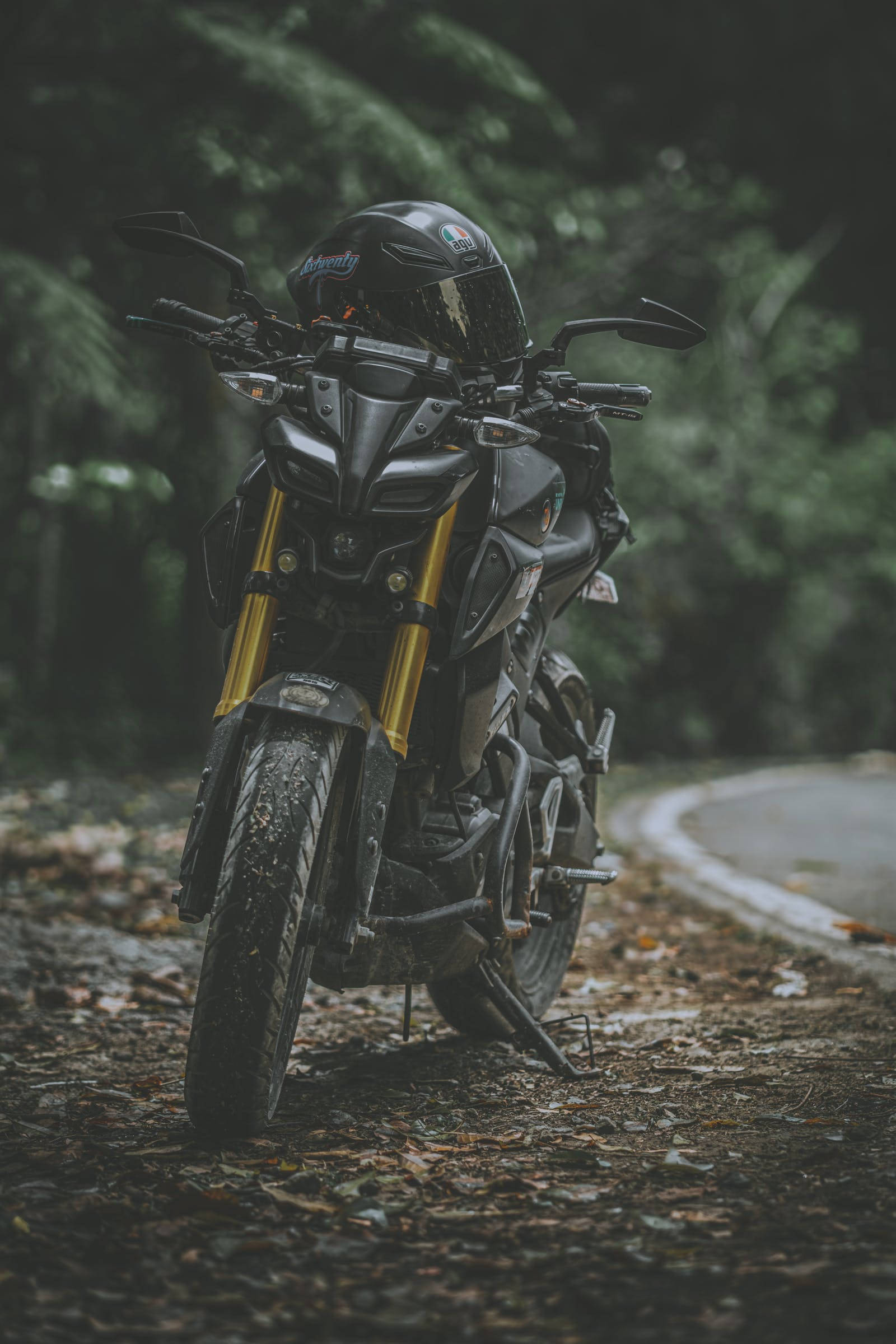 機車 桌布 壁紙 — 一台時尚黑色機車，金色前叉，停泊在綠葉成蔭的林間道路上，安全帽擱在手把上。