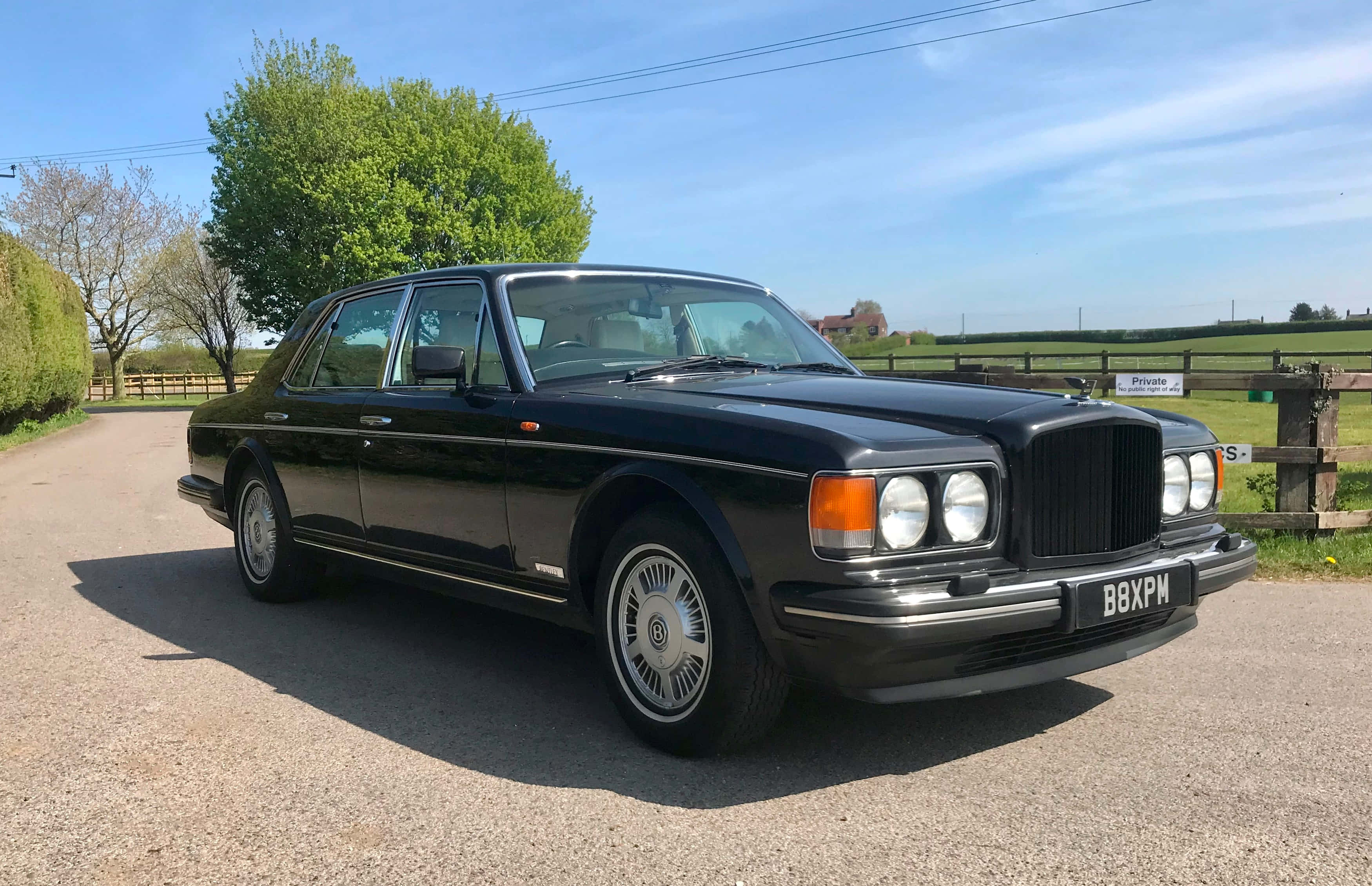 Bentley Eight 桌布 壁紙 — 一台時尚黑色賓利轎車在明亮陽光下閃閃發光，停在碎石車道上，旁邊是翠綠的田野和樹木。