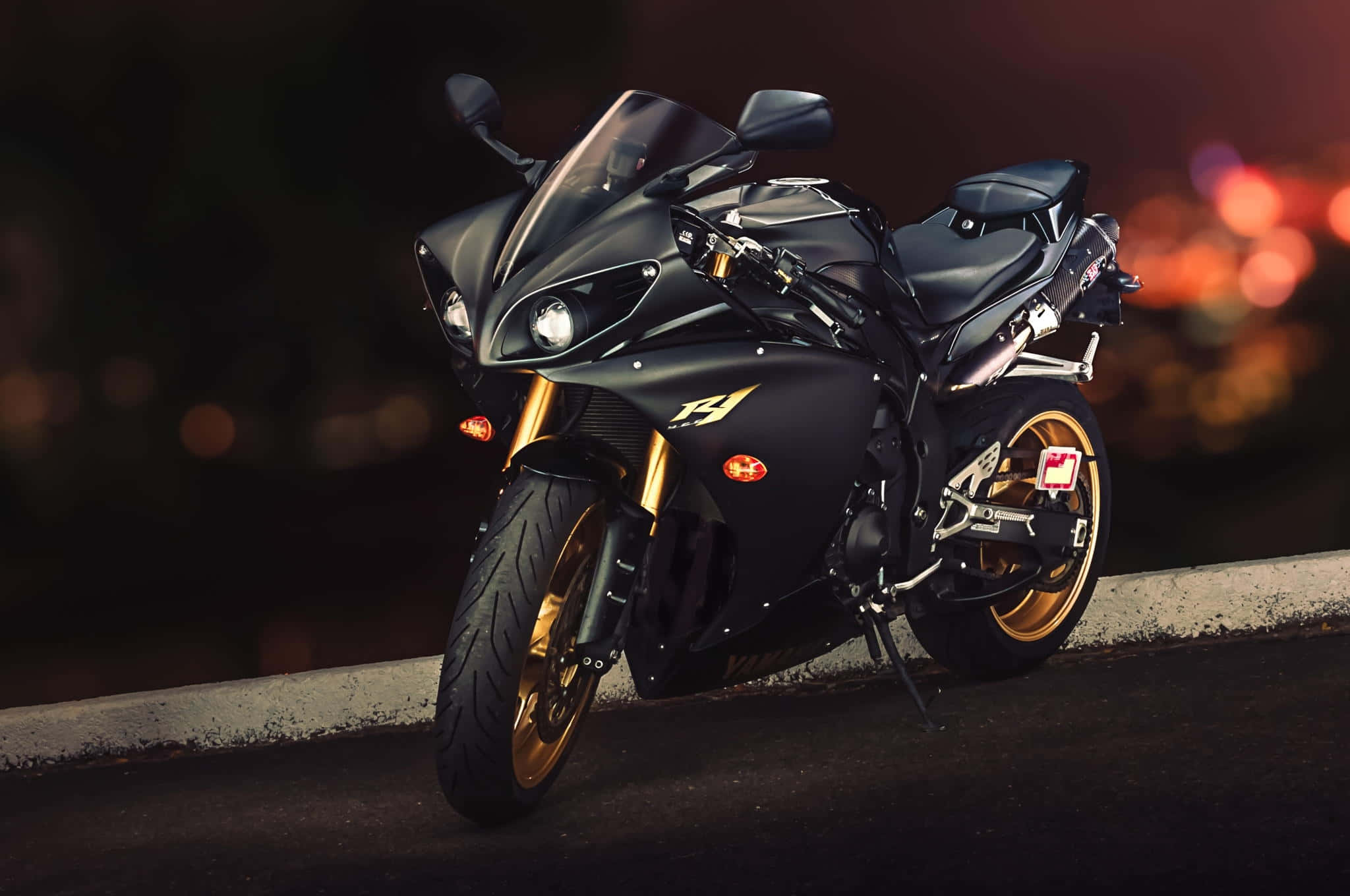 Superbike 桌布 壁紙 — 一台時尚黑色運動機車，帶有金色點綴，夜晚停放在昏暗的道路上，背景是模糊的城市燈光。