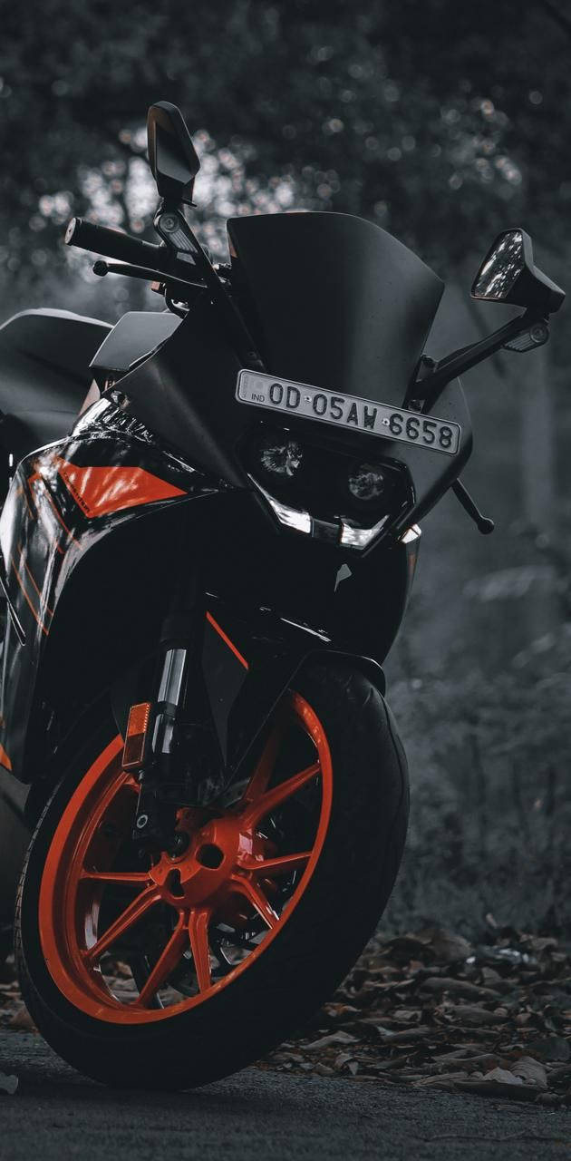 KTM RC 200 桌布 壁紙 — 一台時尚黑色運動機車搭配鮮豔橙色輪胎，停放在樹葉茂密的森林小徑上，燈光營造出氛圍。