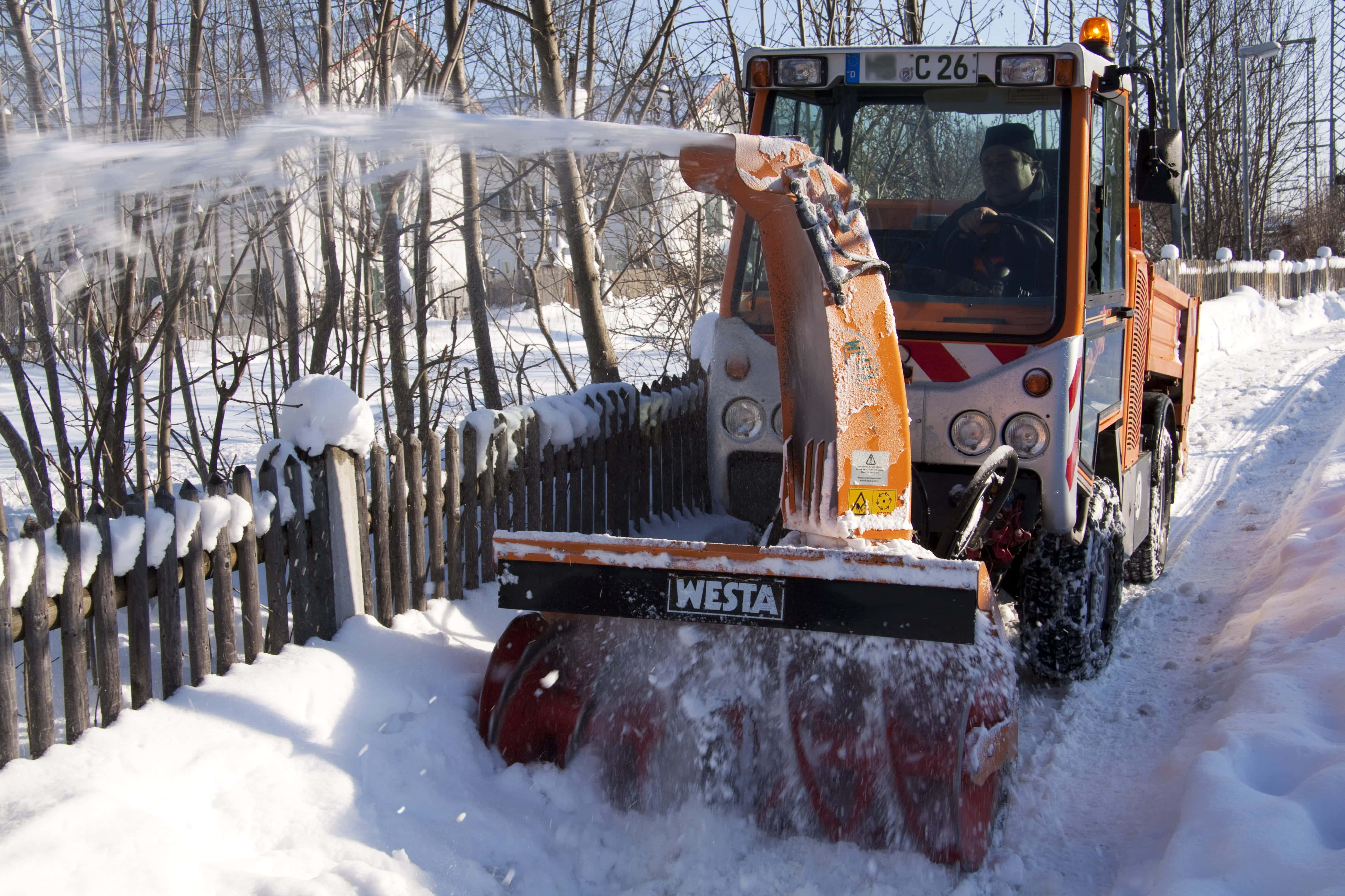 Snowplow 桌布 壁紙 — 一台橙色的吹雪機在明亮的冬日陽光下，清理一條被雪覆蓋的小路，旁邊是一個沾滿雪的木籬笆。