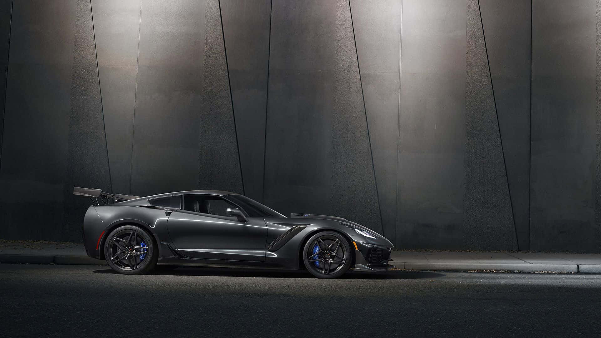 corvette 桌布 壁紙 — 一台流線型消光灰色跑車搭配藍色煞車卡鉗，在戲劇性聚光燈下停泊於深色工業牆前。