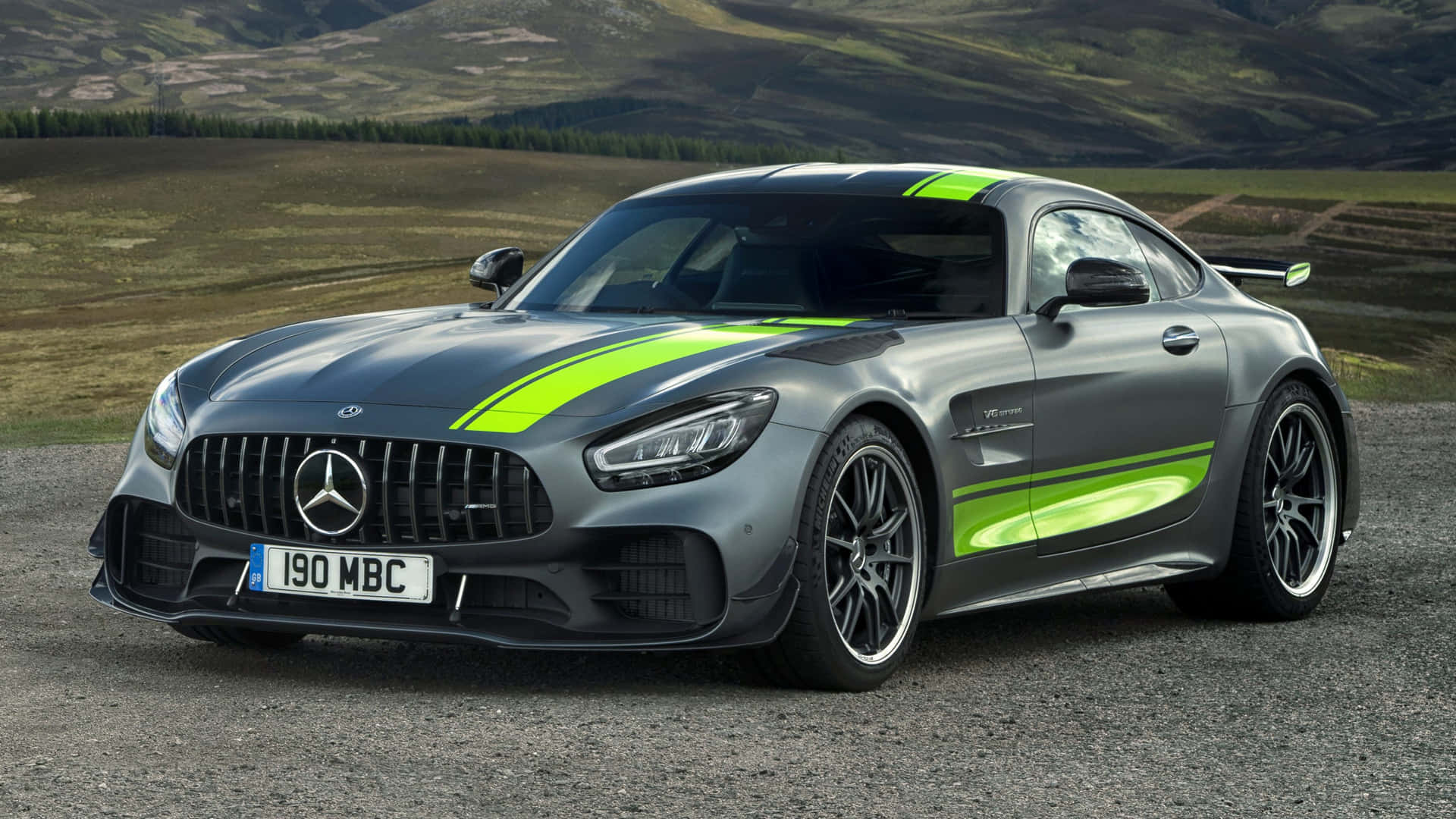 風景 桌布 壁紙 — 一台流線型灰色Mercedes-AMG GT，帶有螢光綠色賽車條紋，停泊在碎石地上，背景是連綿起伏的山丘。