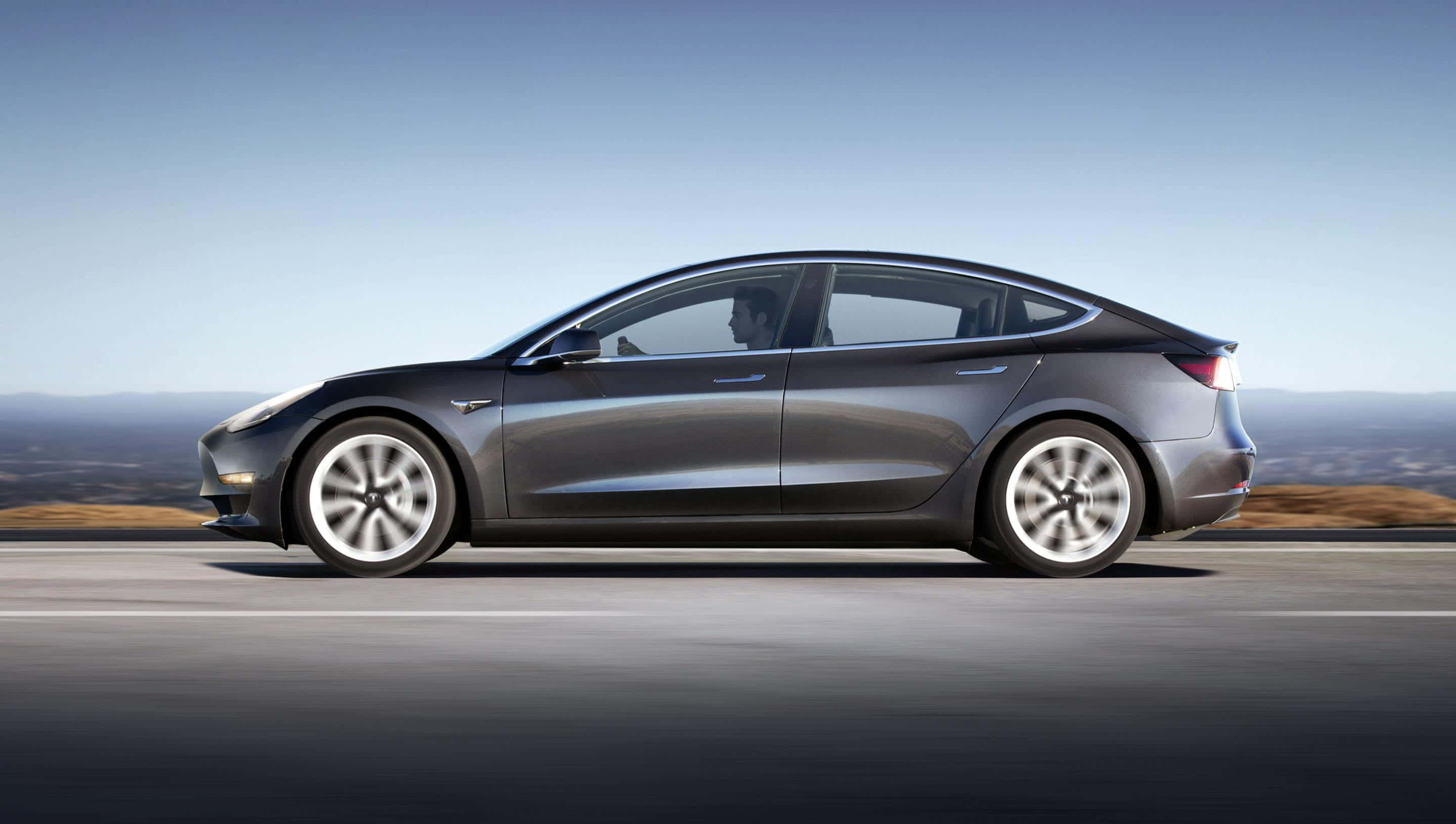 Tesla Model 3 桌布 壁紙 — 一台流線型灰色Tesla Model 3在高速公路上行駛，背景是清澈的藍天和遠山，帶有動態模糊效果。