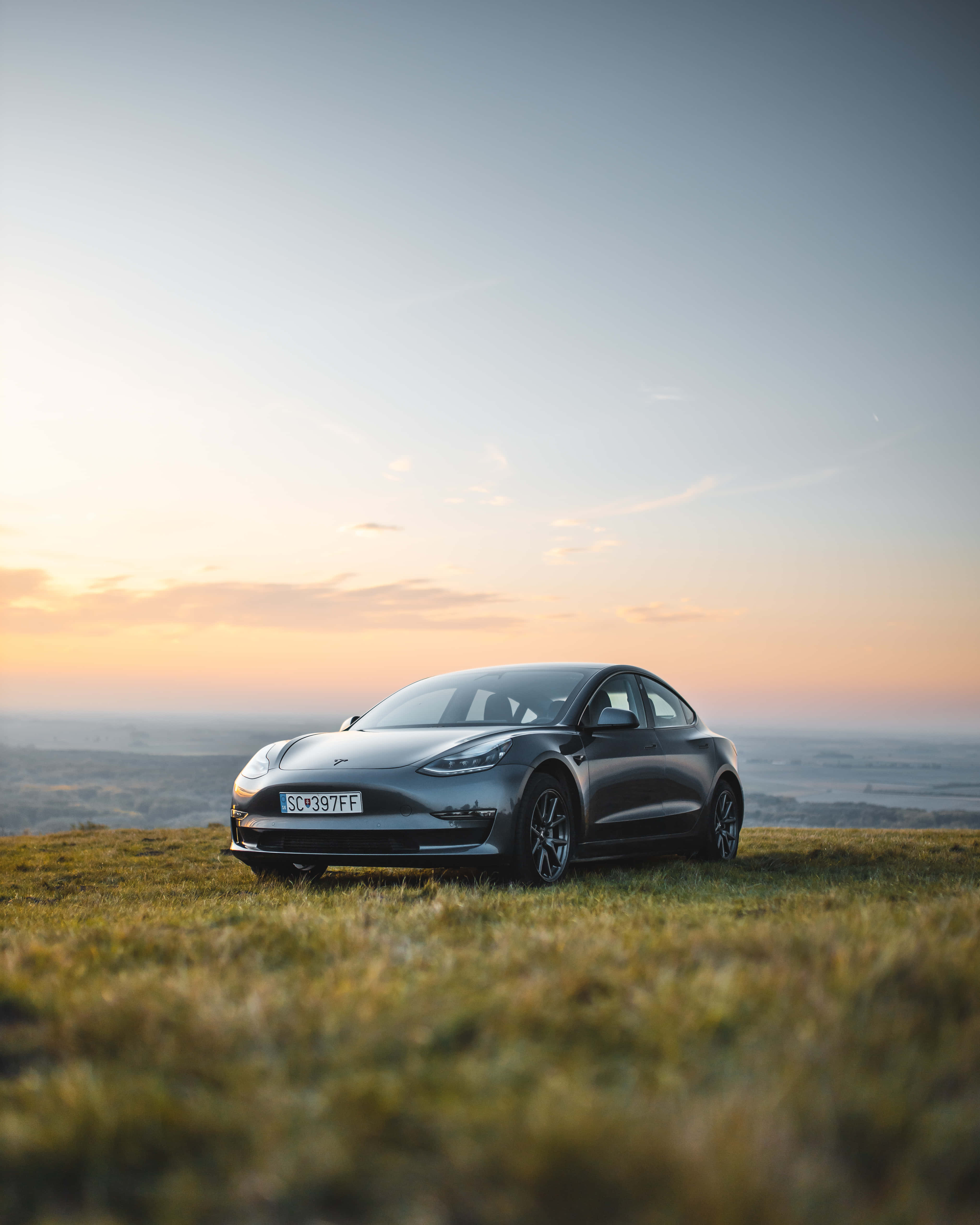 特斯拉 桌布 壁紙 — 一台流線型灰色Tesla Model 3停在日落時分的草地上，上方是柔和的橙色和藍色天空。
