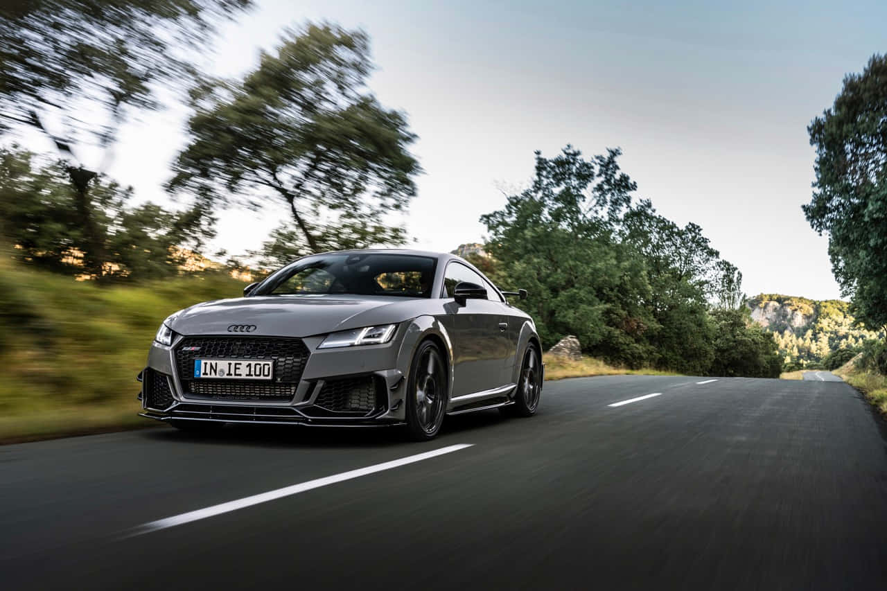 Audi Tt Rs 桌布 壁紙 — 一台流線型灰色奧迪TT RS在蜿蜒的鄉村道路上疾馳，背景是模糊的綠樹和山丘。