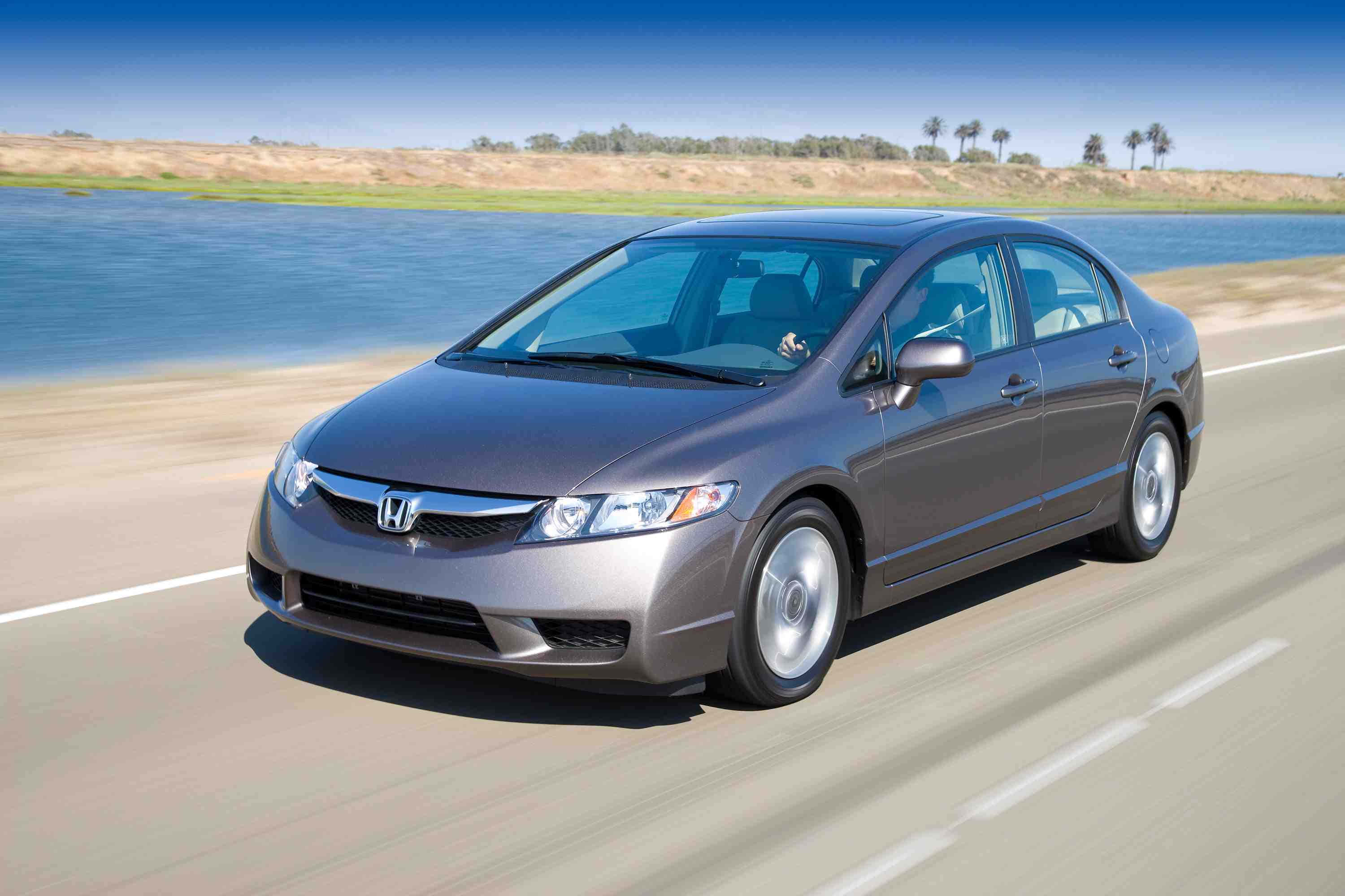 Honda Civic 桌布 壁紙 — 一台流線型灰色本田思域在晴朗天空下，沿著平靜的藍色河流旁陽光普照的高速公路行駛。
