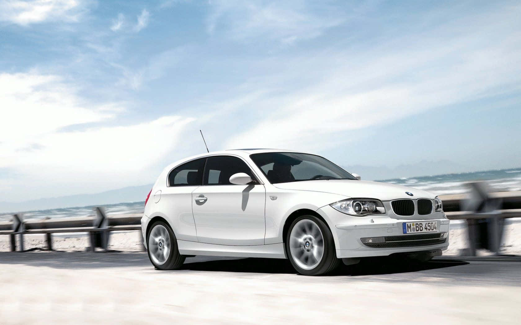 Bmw 1 Series 桌布 壁紙 — 一台流線型白色BMW 1系列掀背車沿著海岸道路行駛，背景是海浪和遠山，天空湛藍明亮。