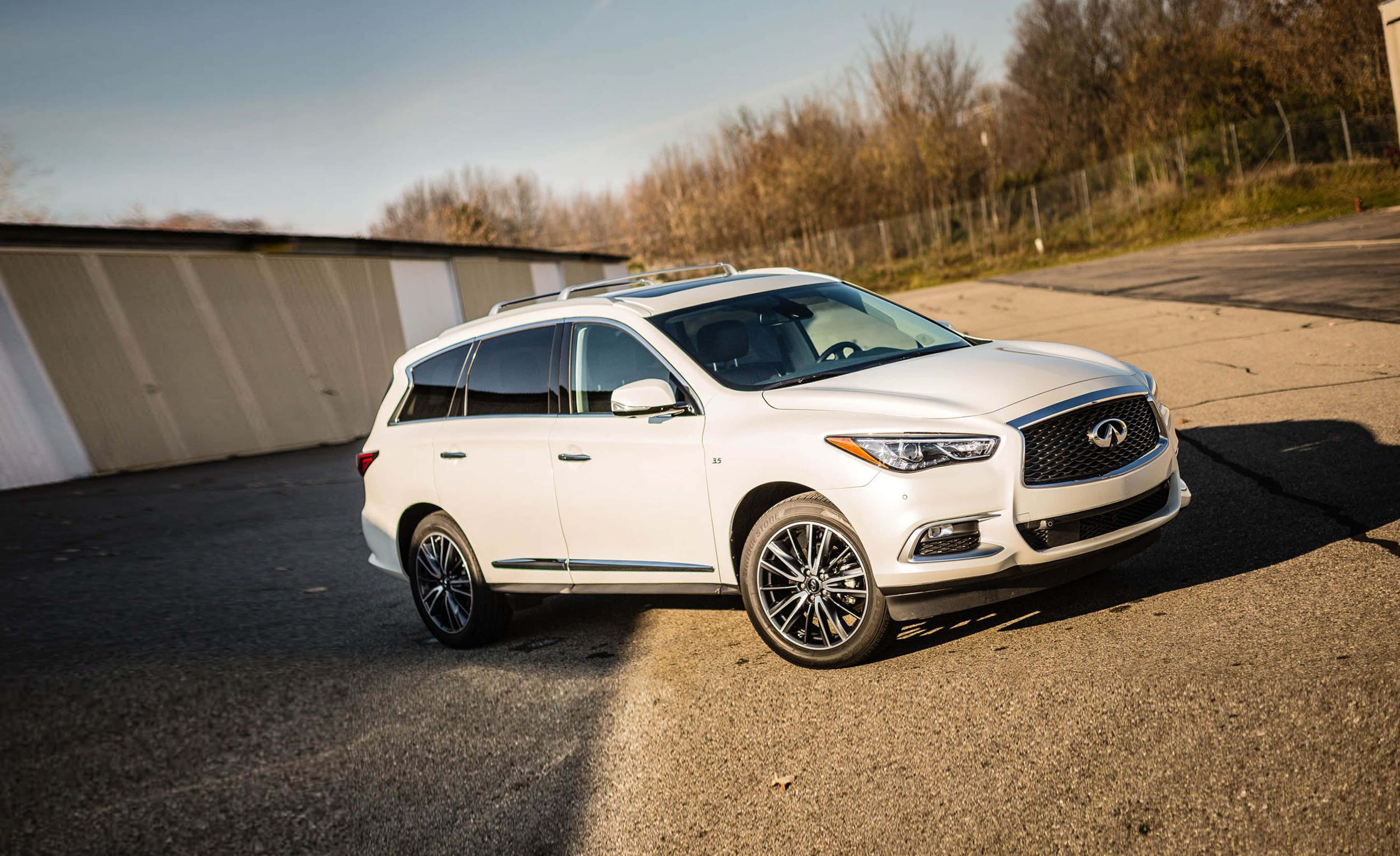 Infiniti Qx60 桌布 壁紙 — 一台流線型白色Infiniti QX60 SUV停放在柏油路上，背景有光禿樹木和倉庫，天空晴朗。