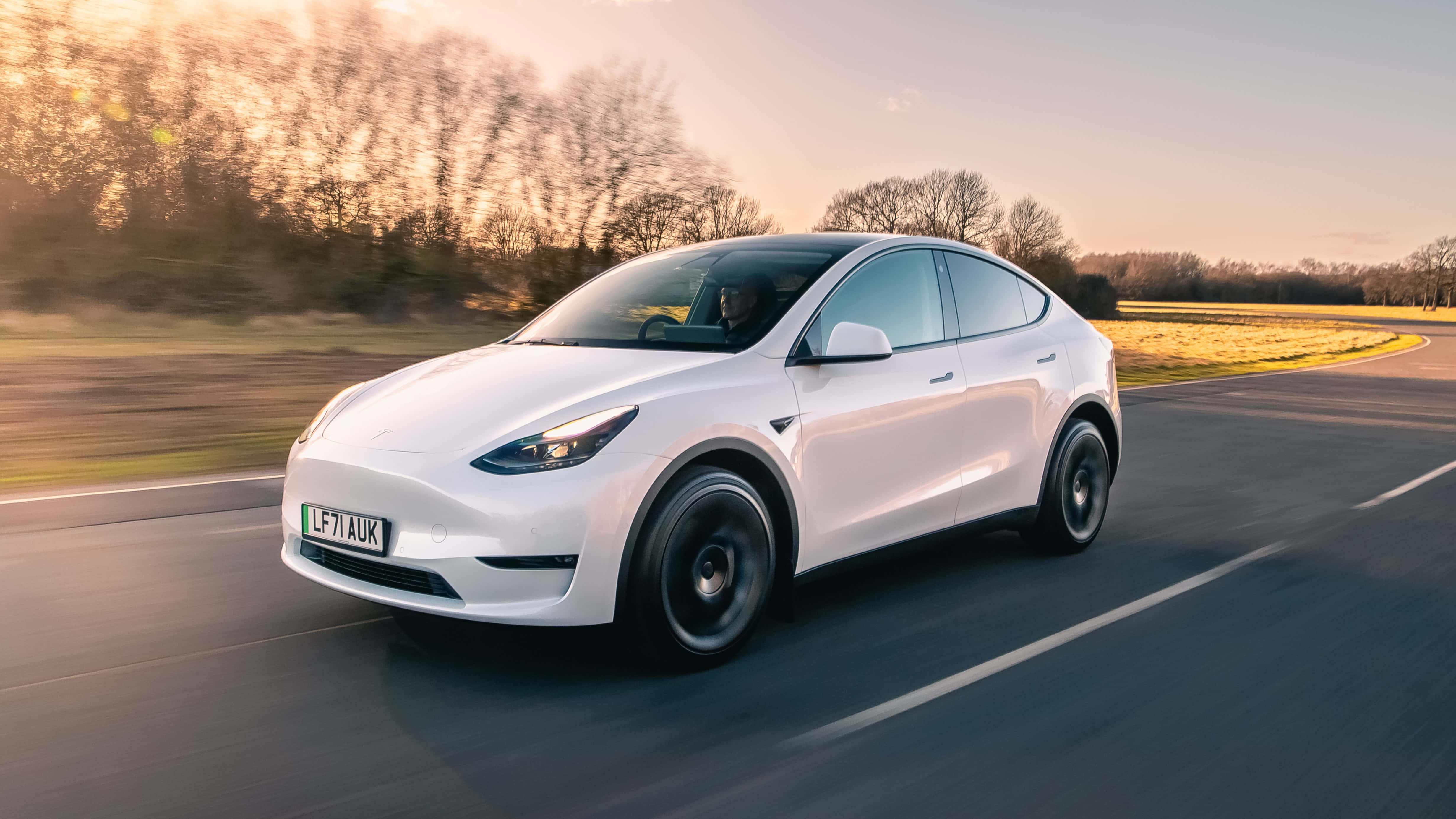 特斯拉 桌布 壁紙 — 一台流線型白色Tesla Model Y在日落時分行駛在蜿蜒的道路上，背景有模糊的樹木和金色的光線。