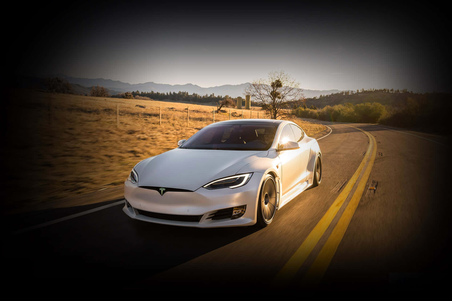 特斯拉 桌布 壁紙 — 一台流線型白色Tesla Model S在黃金時段沿著蜿蜒的鄉村道路行駛，遠處有山巒。