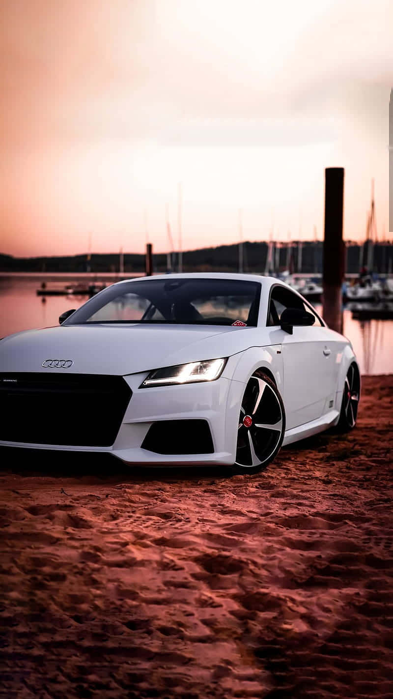 Audi Tt Rs 桌布 壁紙 — 一台流線型白色奧迪跑車停泊在碼頭的微紅沙灘上，背景是日落時分的船隻。