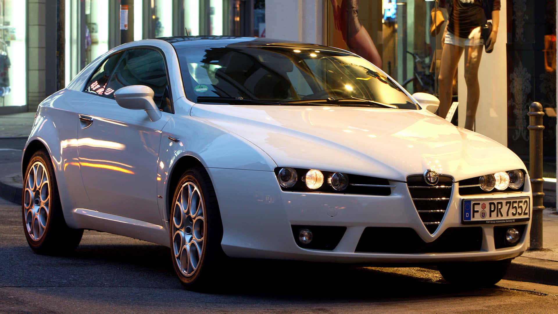 Alfa Romeo Brera 桌布 壁紙 — 一台流線型白色愛快羅密歐跑車在金色街燈下閃閃發光，停放在商店櫥窗附近的城市人行道上。
