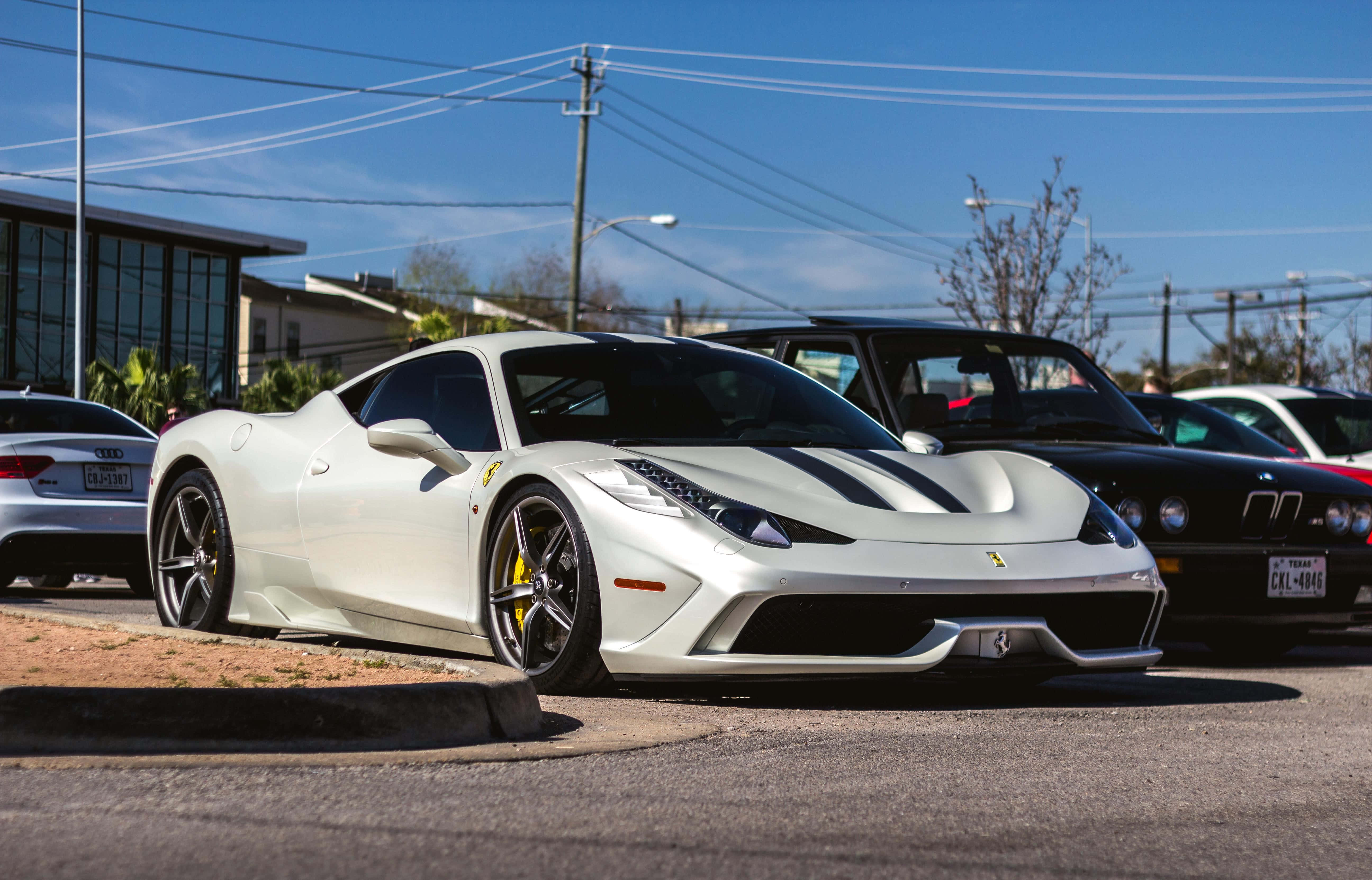 Ferrari 458 Speciale 桌布 壁紙 — 一台流線型白色法拉利跑車帶有黑色賽車條紋，停放在陽光明媚的城市地段，旁邊有其他豪華車輛。