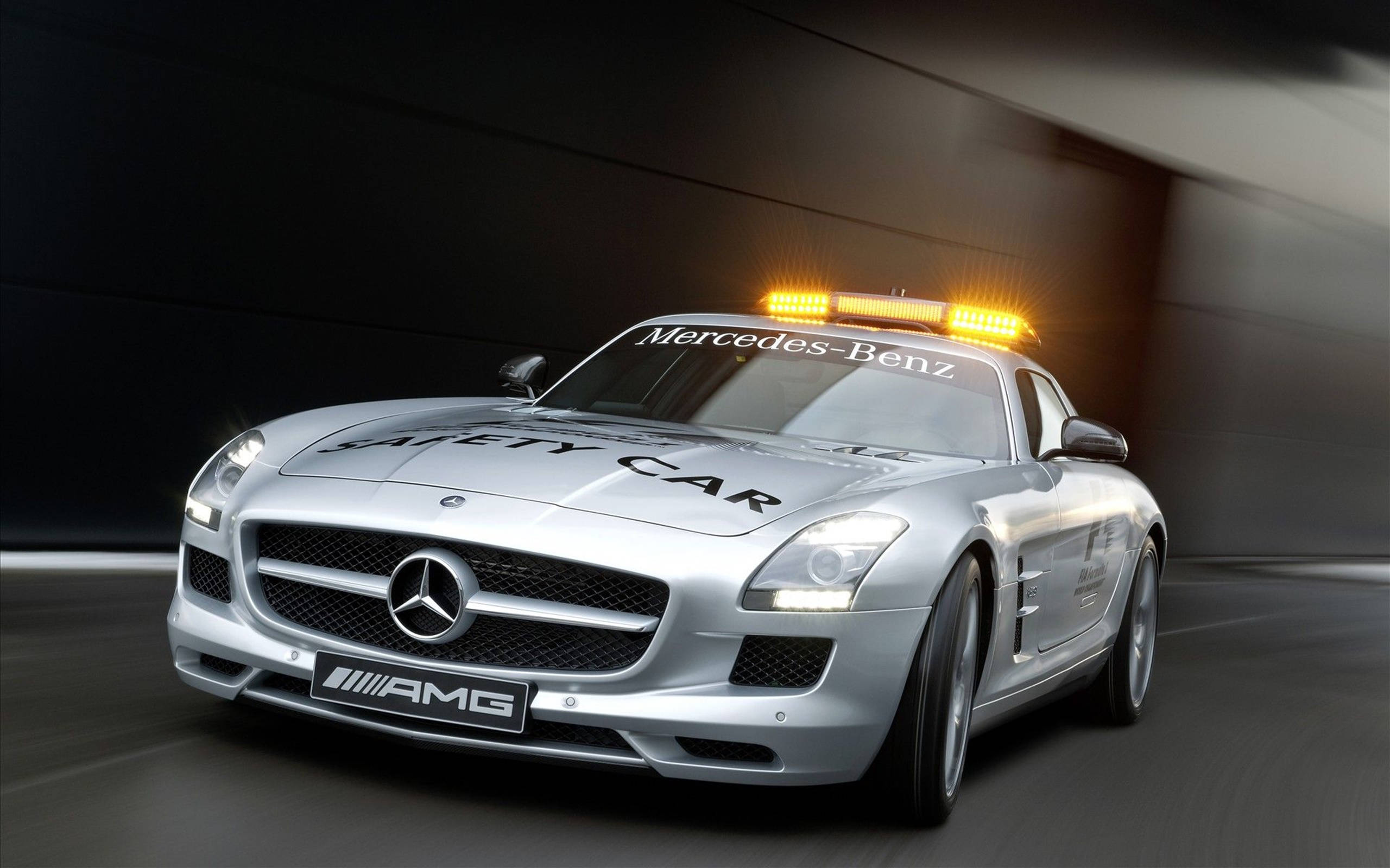 高清 桌布 壁紙 — 一台流線型的銀色 Mercedes-Benz SLS AMG 安全車在昏暗的隧道中疾馳，隧道頂部散發著琥珀色的燈光。