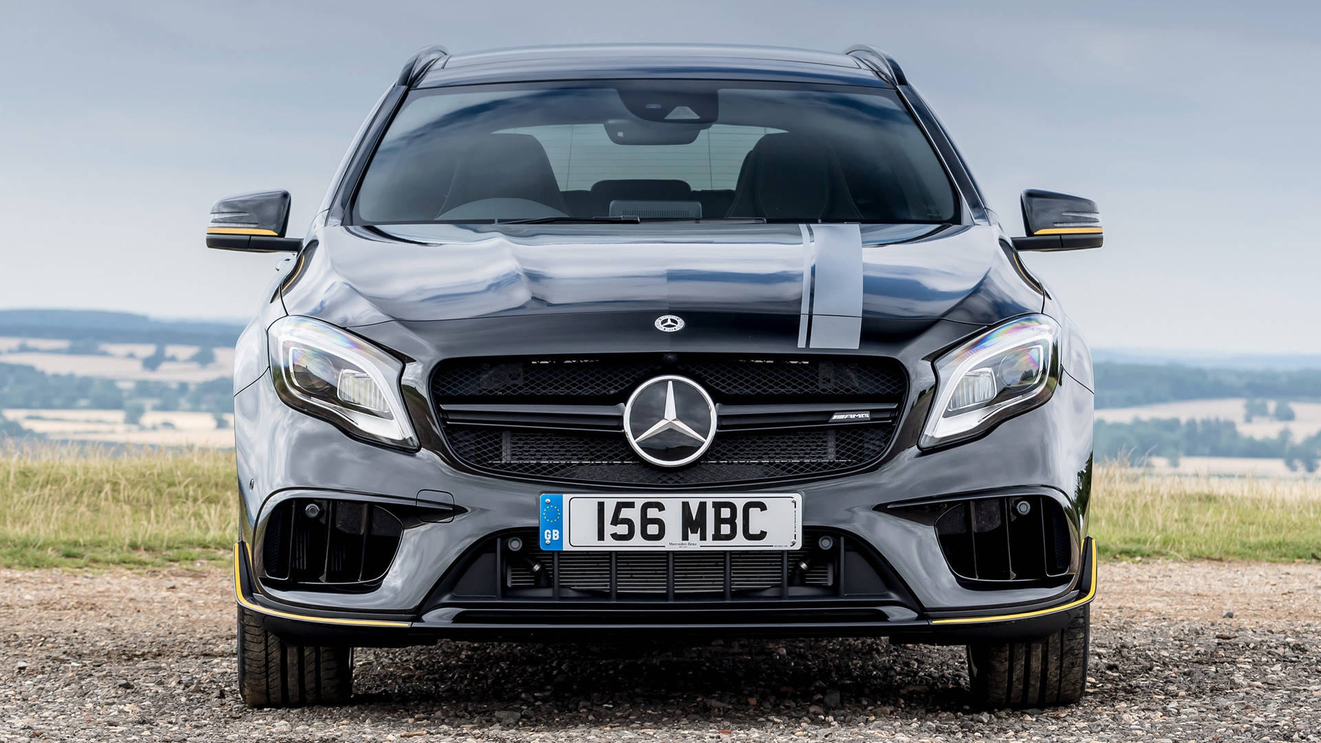 amg iPhone 桌布 壁紙 — 一台流線型的銀色 Mercedes-AMG A45 S 停在碎石路上，背後是連綿起伏的綠色山丘，天空柔和湛藍。
