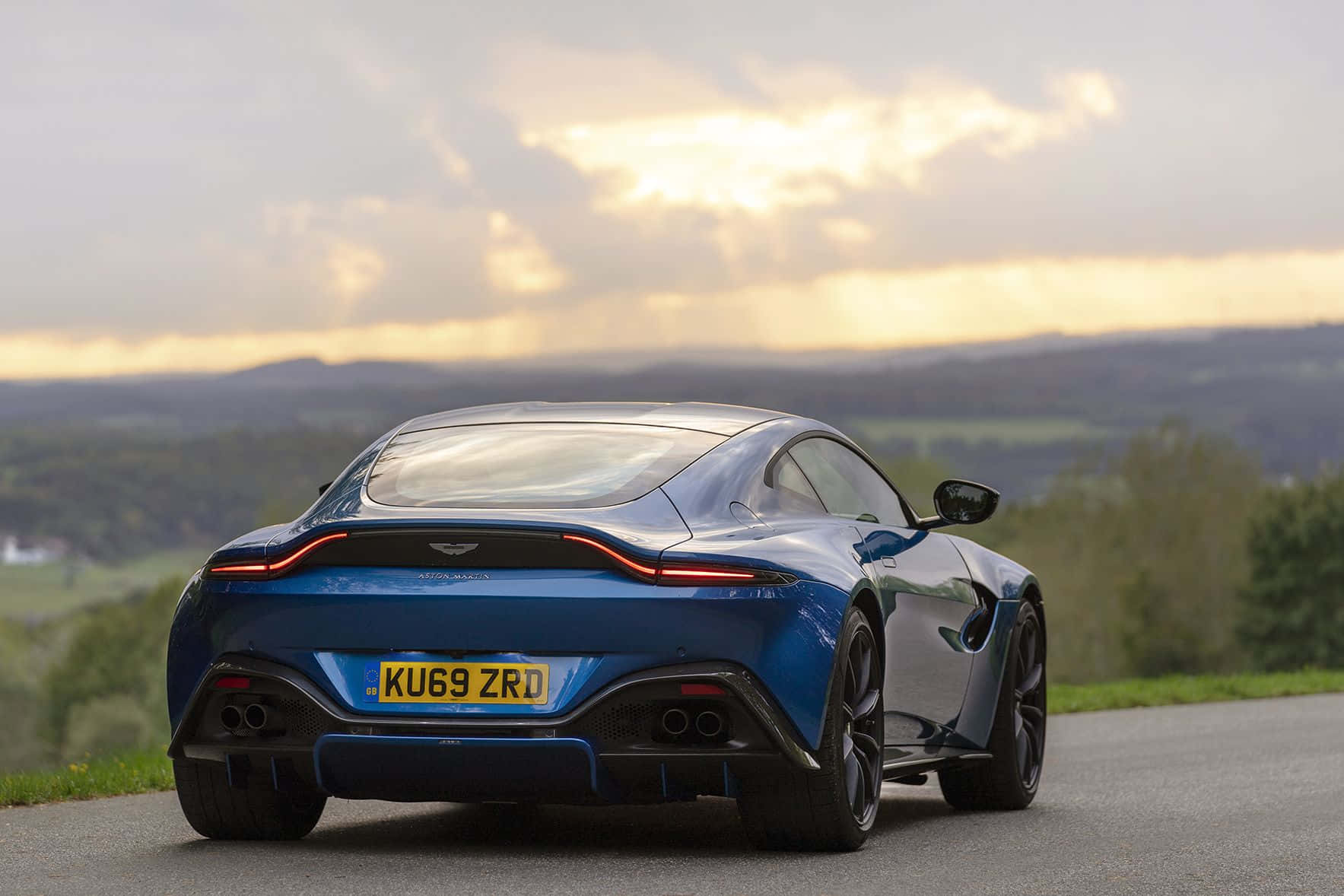 奧斯頓馬丁 桌布 壁紙 — 一台流線型藍色Aston Martin Vantage跑車在日落時分沿著蜿蜒的道路行駛，背景是起伏的丘陵。