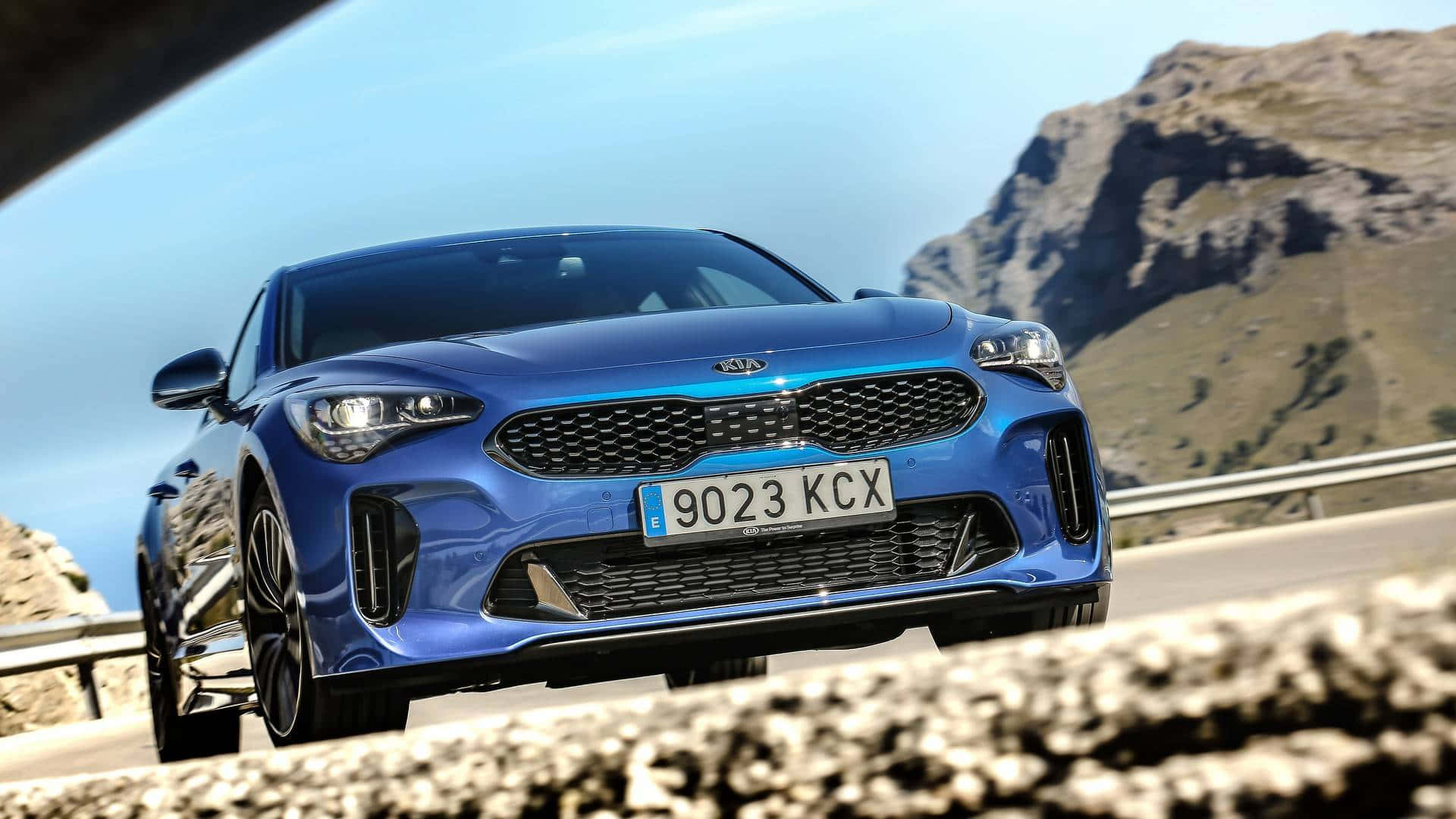 Kia Stinger 桌布 壁紙 — 一台流線型藍色Kia Stinger跑車在晴朗天空下行駛於山路上，背景是崎嶇的山峰。