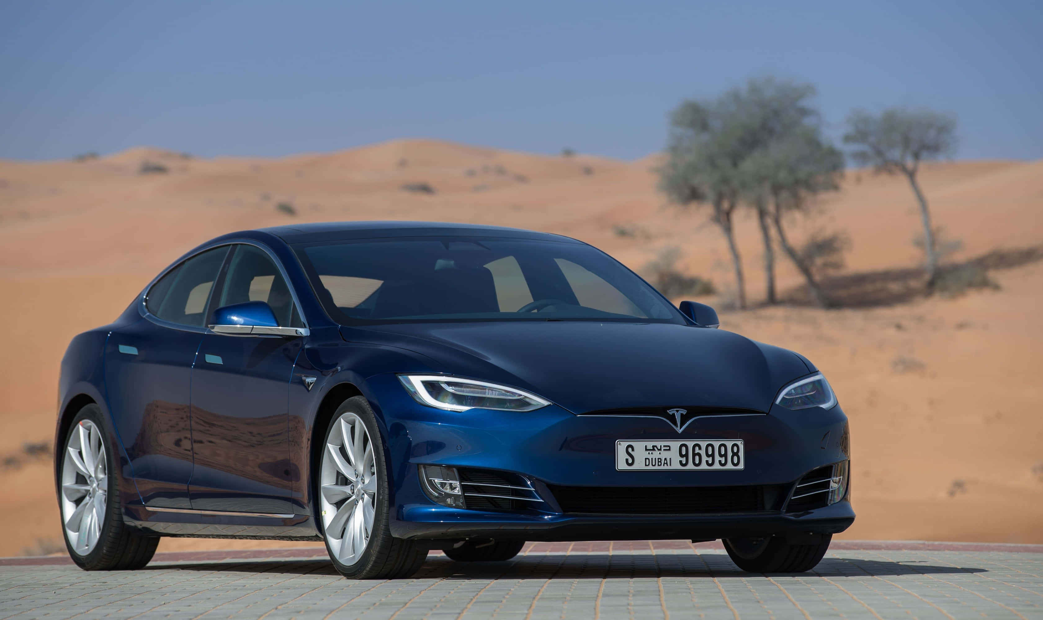 特斯拉 桌布 壁紙 — 一台流線型藍色Tesla Model S停放在鋪砌路面上，背景是金色沙漠沙丘和稀疏樹木，天空清澈湛藍。