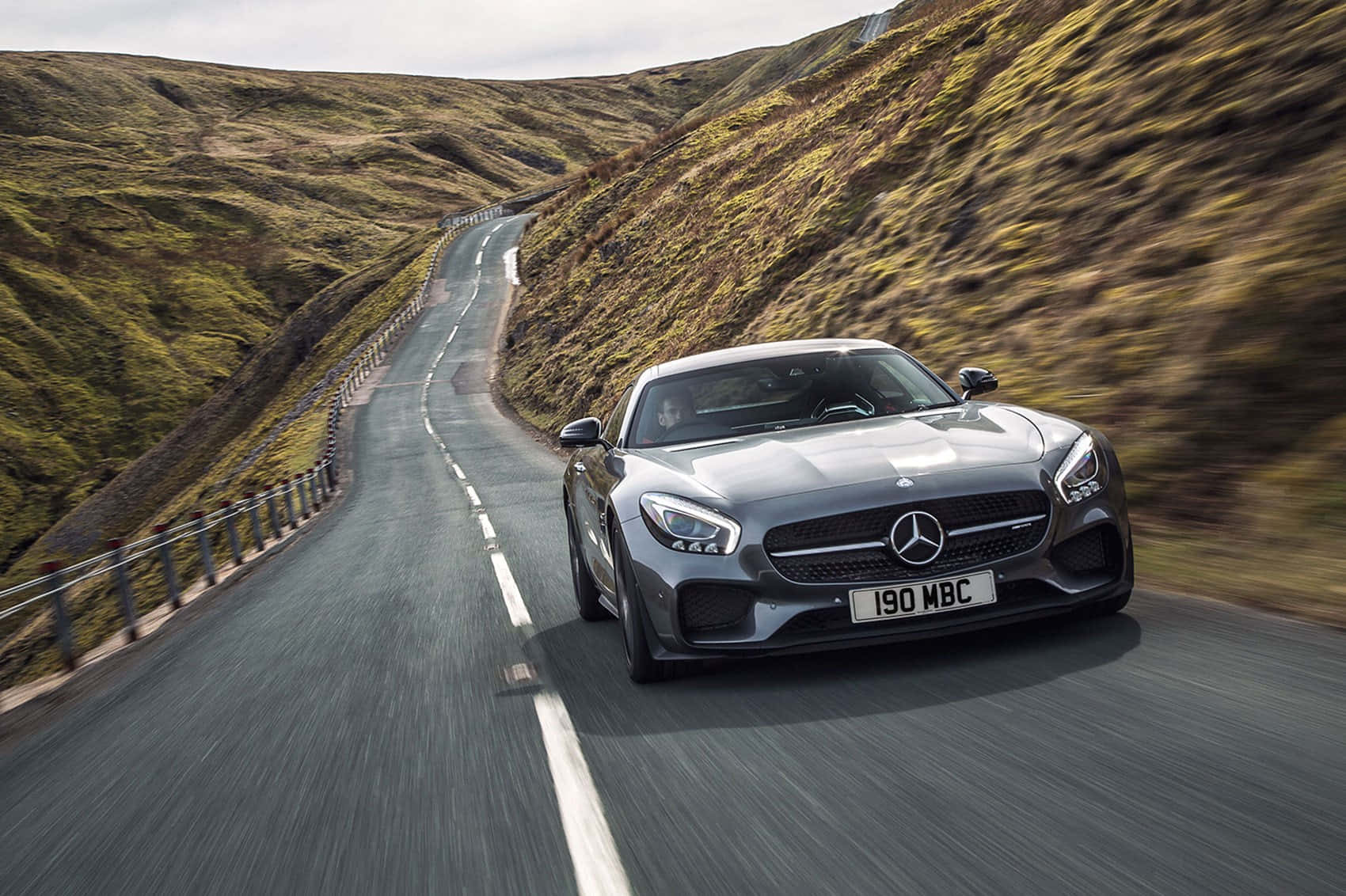 汽車 桌布 壁紙 — 一台流線型銀色 Mercedes-AMG GT 在陰天之下，沿著蜿蜒的山路穿過翠綠的山丘奔馳。