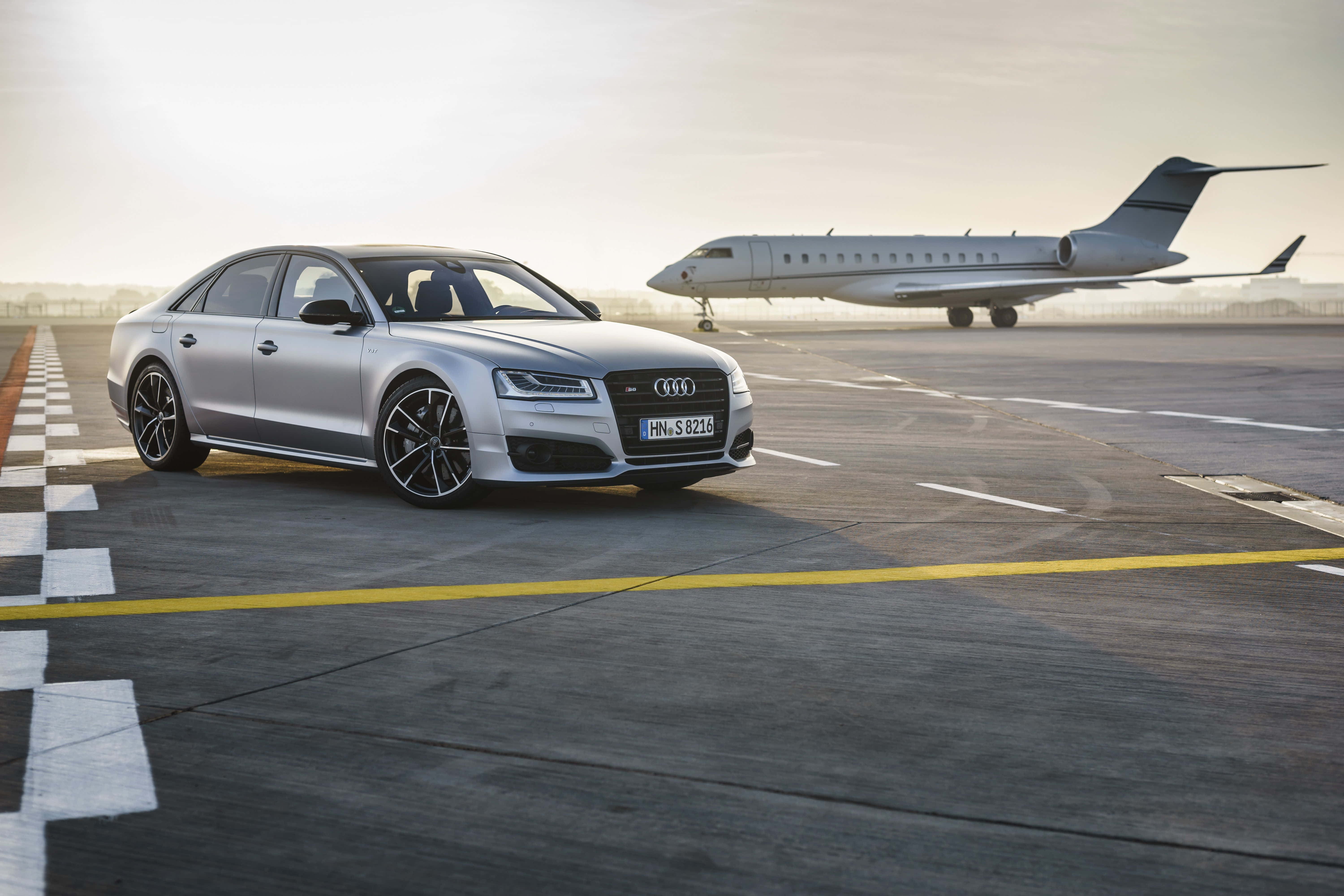 Audi S8 桌布 壁紙 — 一台流線型銀色奧迪S8轎車停在機場停機坪上，背景有私人飛機，沐浴在柔和的金色光線中。