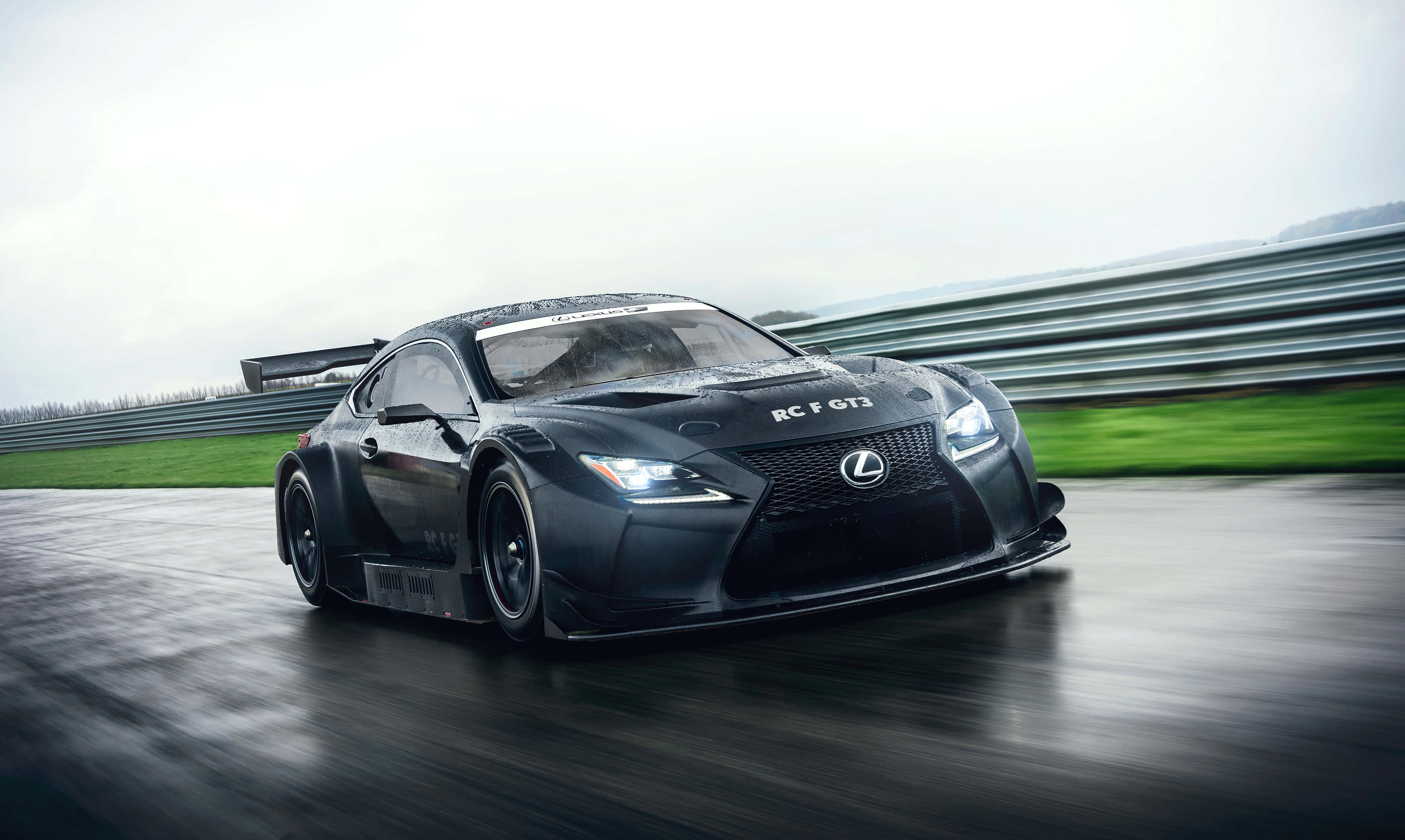 Lexus Rc F 桌布 壁紙 — 一台流線型黑色 Lexus RC F GT3 賽車在潮濕的賽道上疾馳，背景是模糊的綠色草地和護欄。