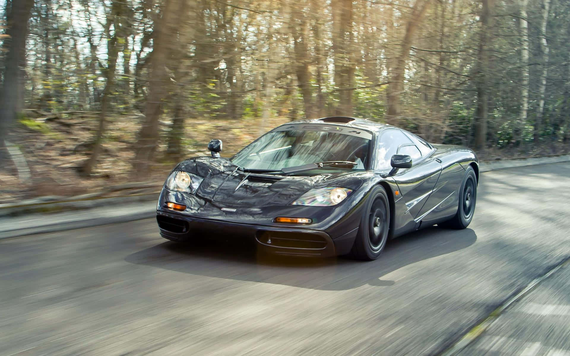 Mclaren F1 桌布 壁紙 — 一台流線型黑色McLaren F1在陽光明媚的森林中疾馳，道路和樹木呈現動態模糊效果。