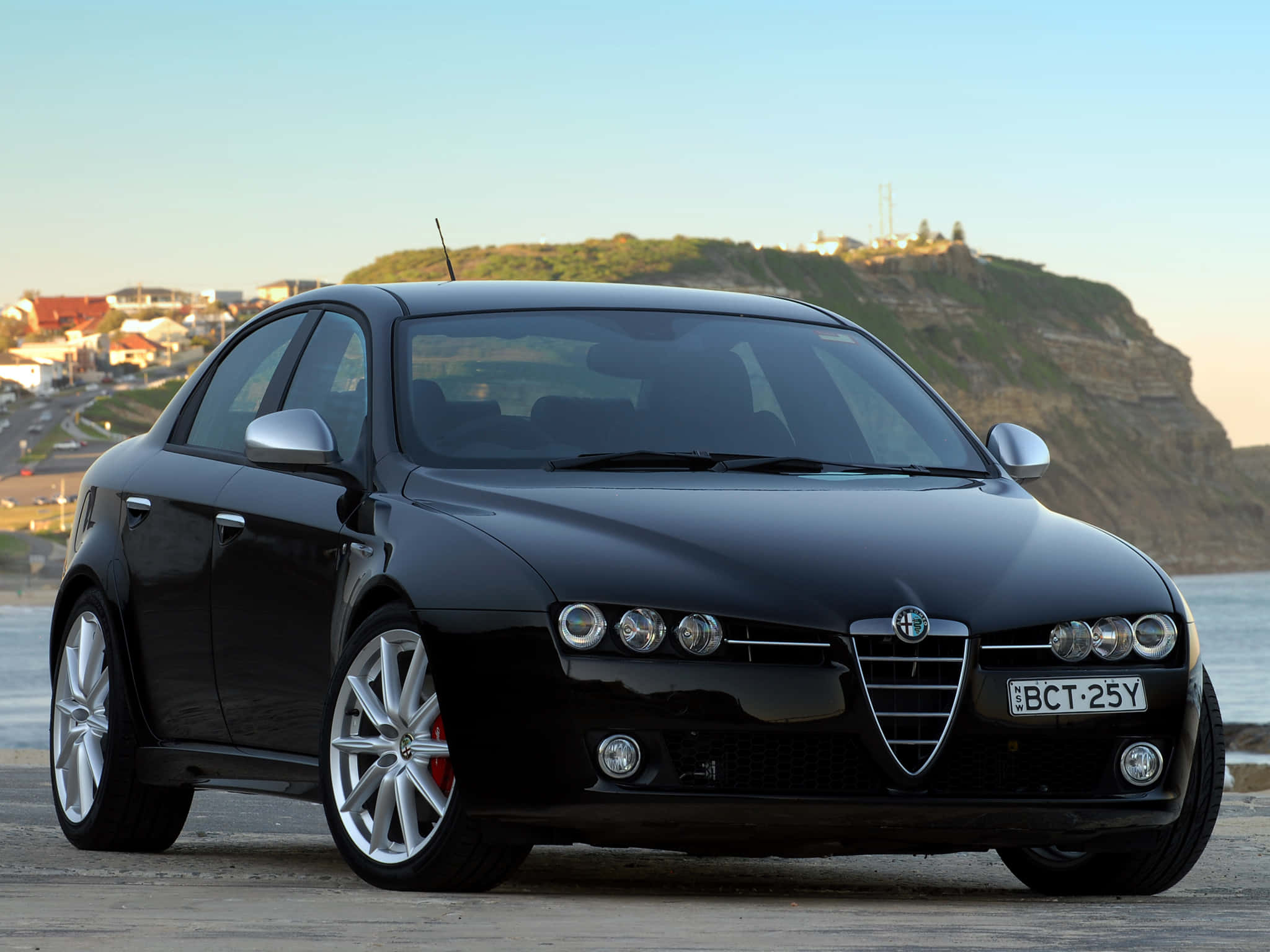 Alfa Romeo 159 桌布 壁紙 — 一台流線型黑色愛快羅密歐轎車停泊在沿海公路上，背景是懸崖小鎮和海洋。