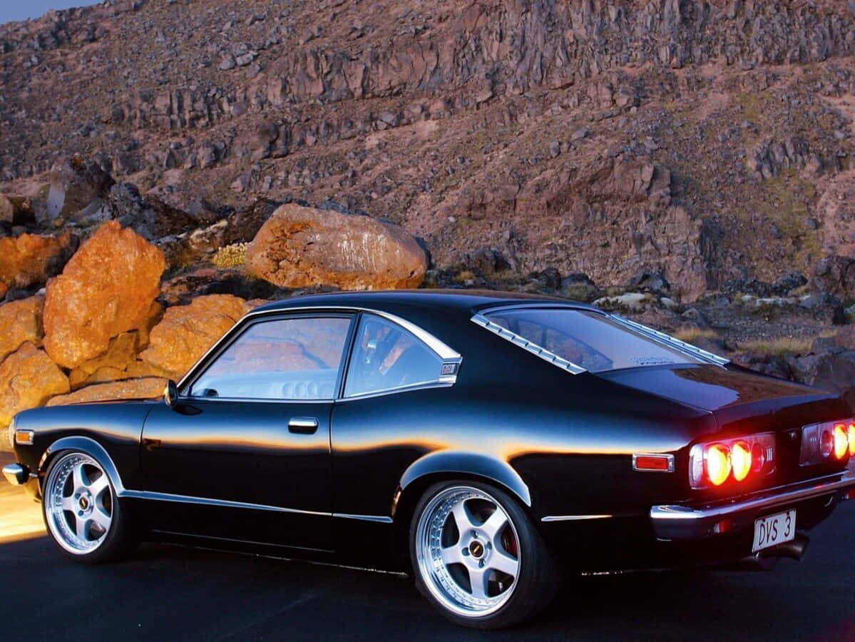 Mazda Rx-3 桌布 壁紙 — 一台流線型黑色經典跑車，帶有發光的紅色尾燈，停在沙漠公路上，旁邊是崎嶇的橙色和棕色岩層。