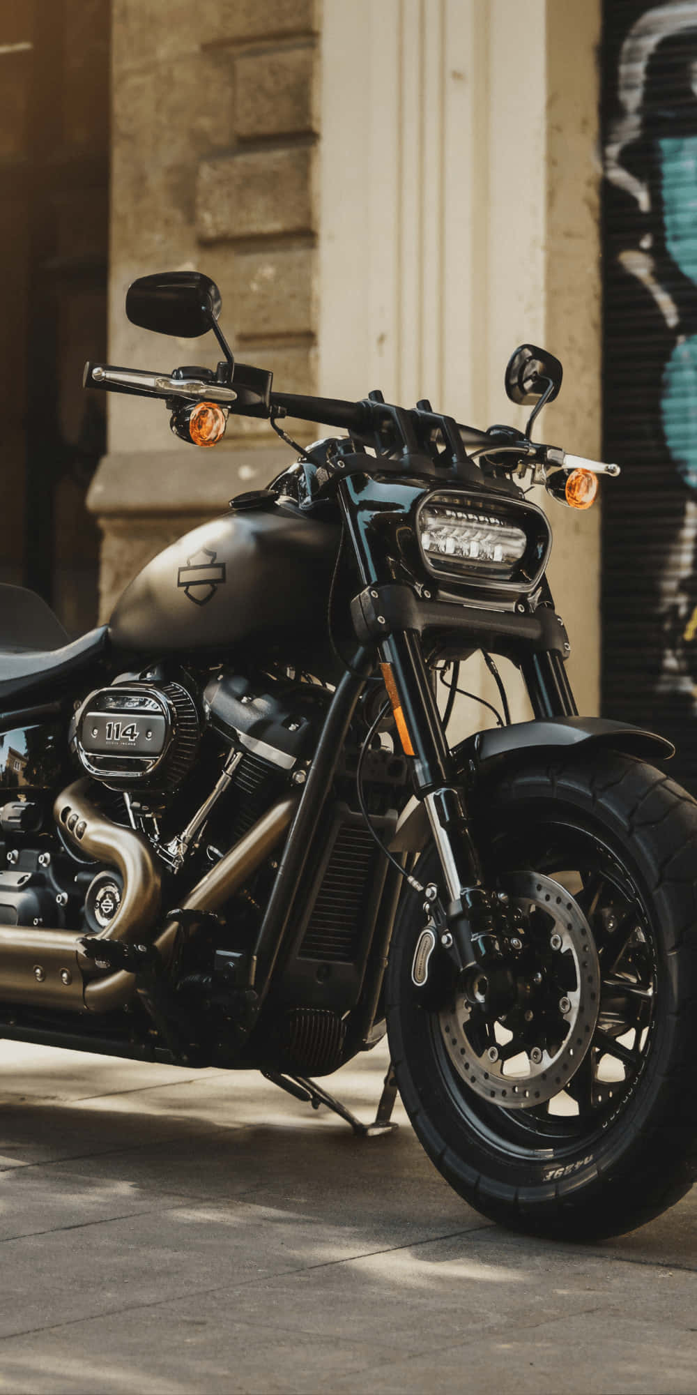 Harley Davidson HD 桌布 壁紙 — 一台消光黑色哈雷戴維森機車停在陽光明媚的城市人行道上，旁邊是佈滿塗鴉的牆壁。