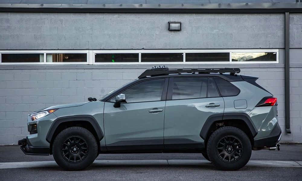 Family Car 桌布 壁紙 — 一台消光鼠尾草綠色豐田RAV4，搭配黑色飾條和車頂行李架，停在灰色磚造建築前。