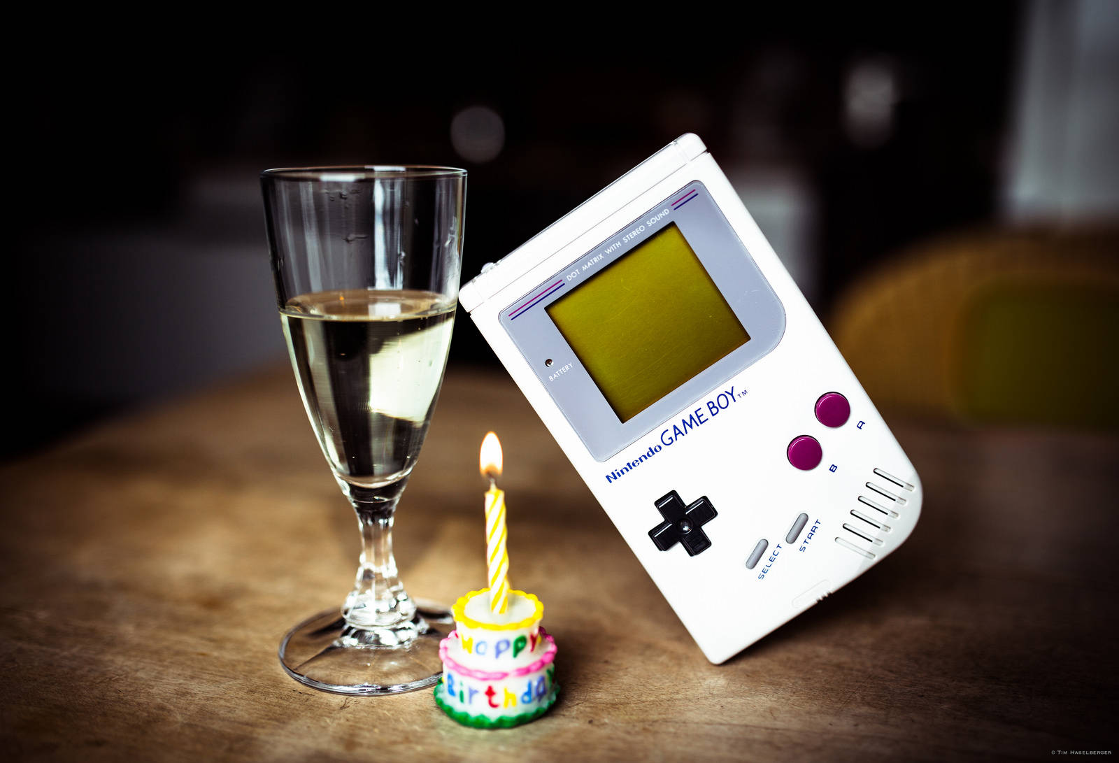 game 桌布 壁紙 — 一台白色 Game Boy 靠在木桌上香檳杯和插著一支點燃蠟燭的小生日蛋糕旁邊。