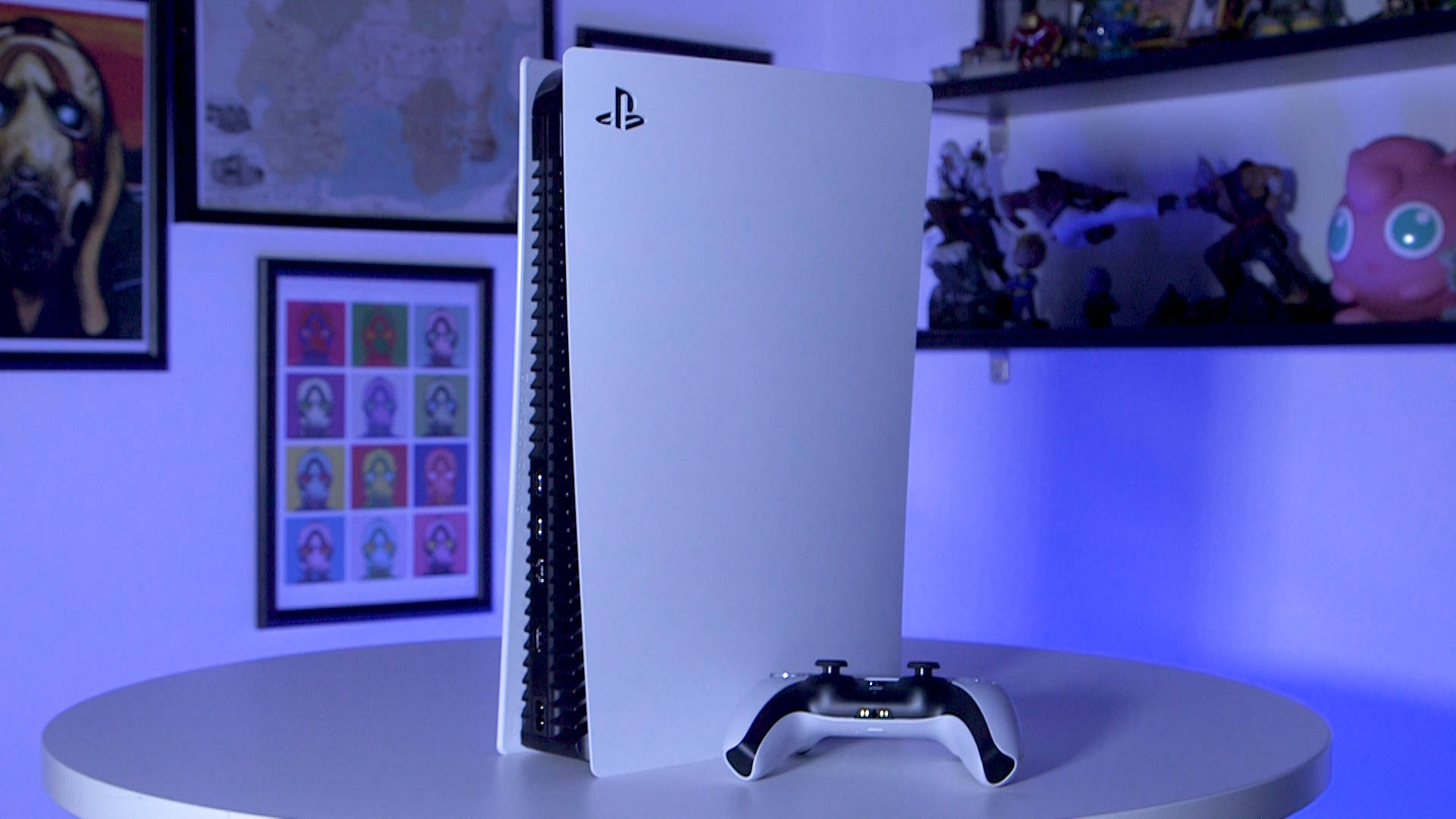 PS5 動態圖片 桌布 壁紙 — 一台白色PlayStation 5主機和控制器放在圓桌上，背後牆上有藍色環境燈光以及普普藝術和遊戲海報。