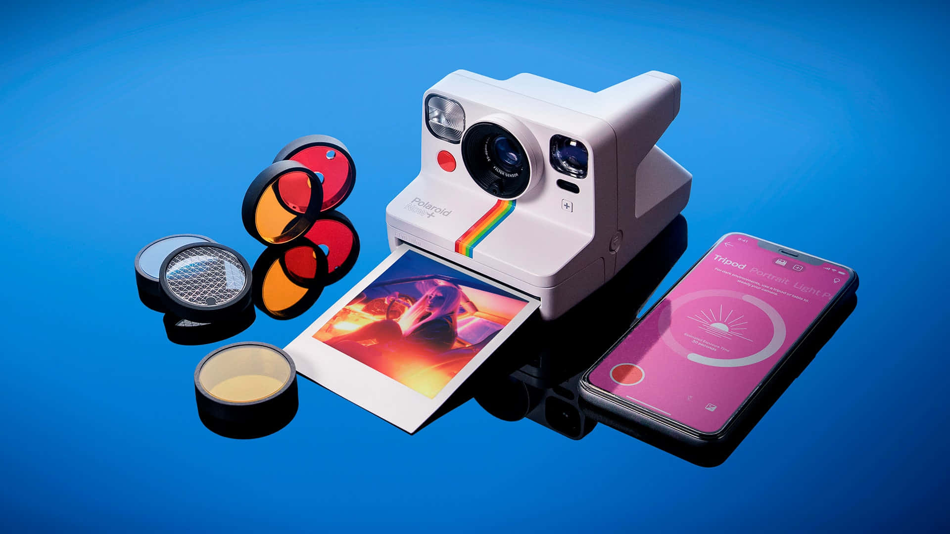 手機 桌布 壁紙 — 一台白色Polaroid相機在亮面藍色表面上，於彩色濾鏡和智慧型手機旁邊彈出一張鮮豔的照片。