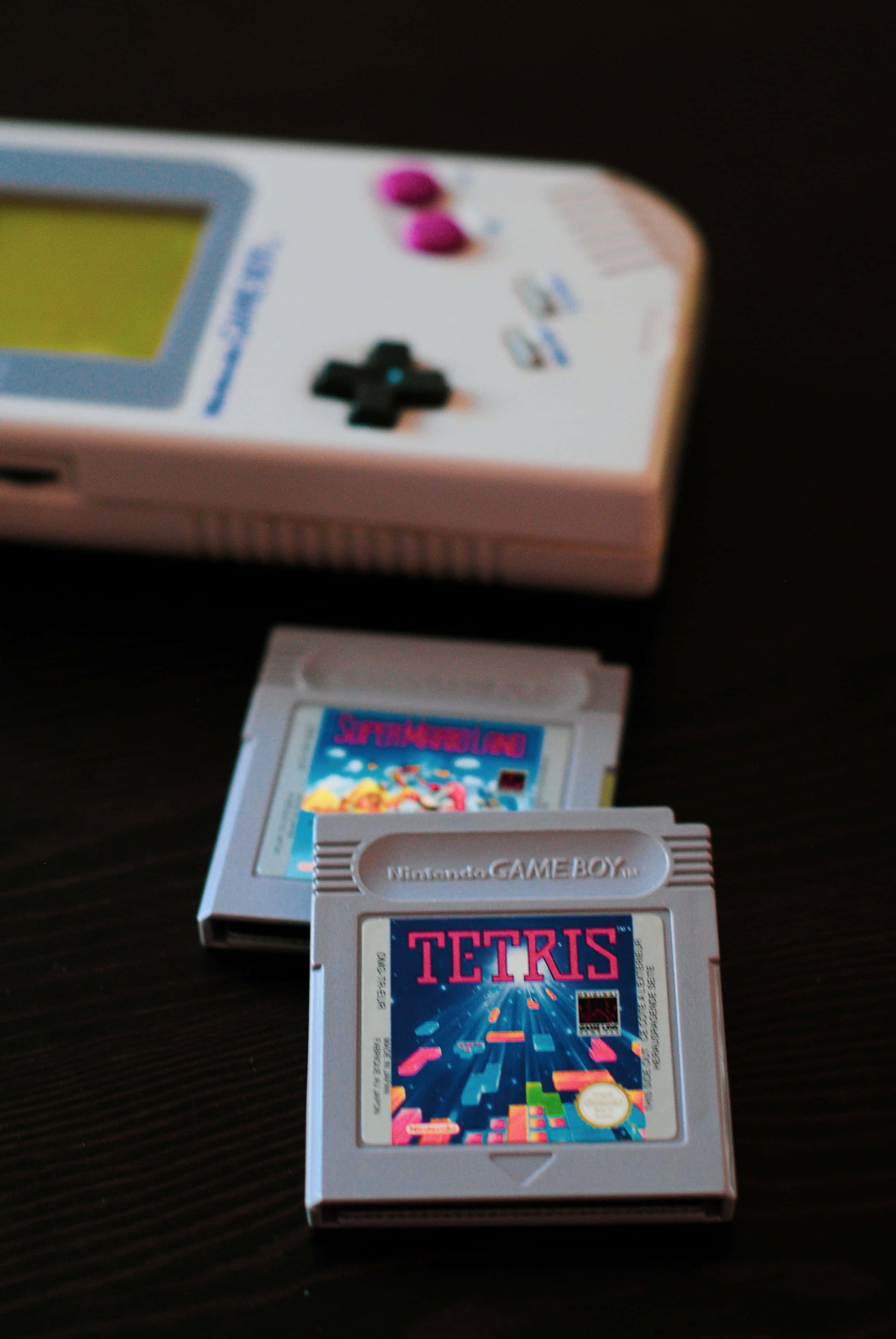 Gaming Consoles 桌布 壁紙 — 一台白色任天堂Game Boy，上面有兩張遊戲卡帶俄羅斯方塊和超級瑪利歐樂園，放在深色木質表面上。