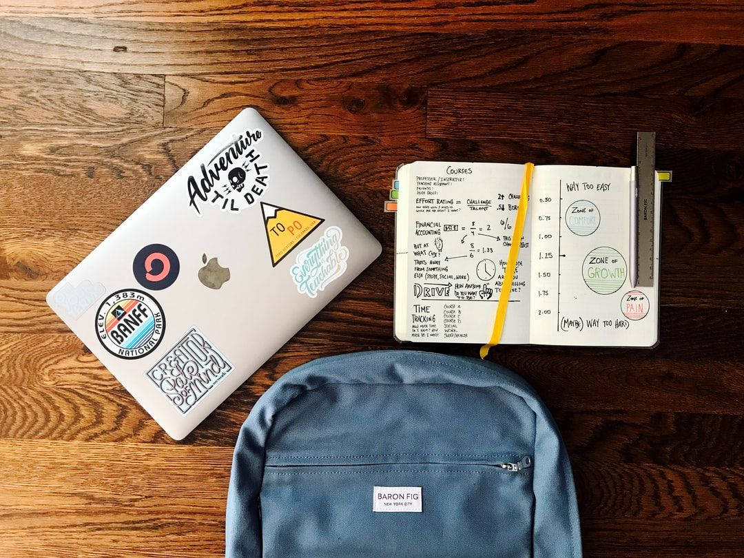 Studying 桌布 壁紙 — 一台白色筆記型電腦上貼滿彩色貼紙，旁邊放著一本打開的筆記本，上面有手寫目標，還有一個藍色背包，放在豐富的木地板上。
