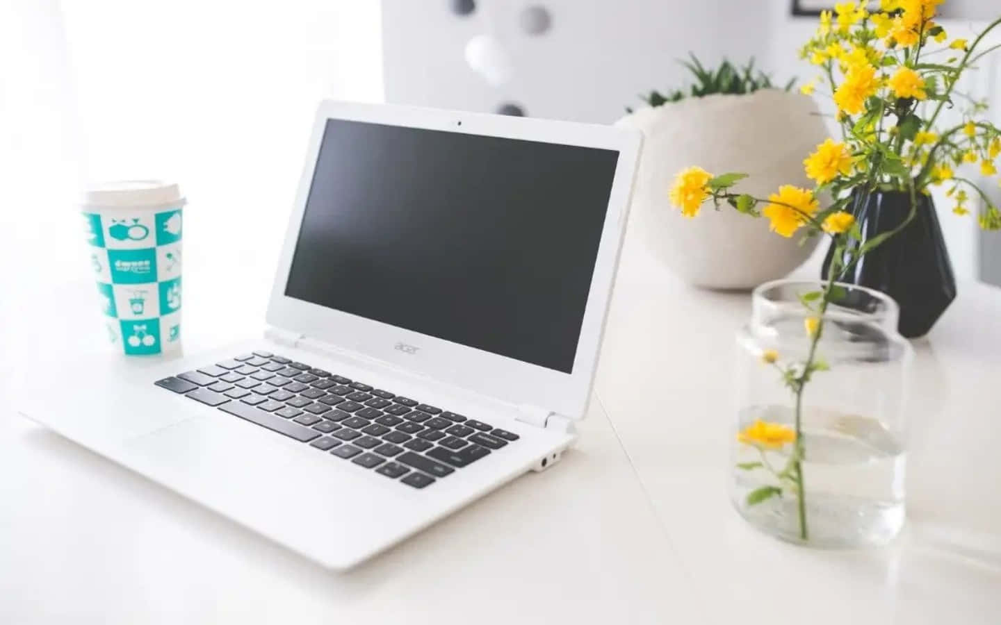 Chromebook 桌布 壁紙 — 一台白色筆記型電腦放在明亮的白色桌子上，旁邊有一個青綠色的咖啡杯和玻璃及陶瓷花瓶中的黃色花朵。