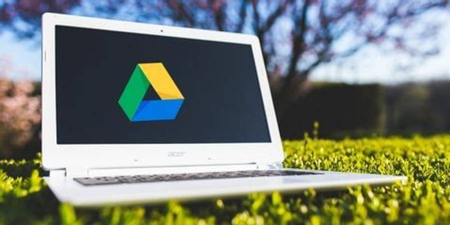 Google Drive 桌布 壁紙 — 一台白色筆記型電腦顯示Google Drive標誌，打開放在鮮綠色草地上，背景是明亮的藍天和模糊的樹木。