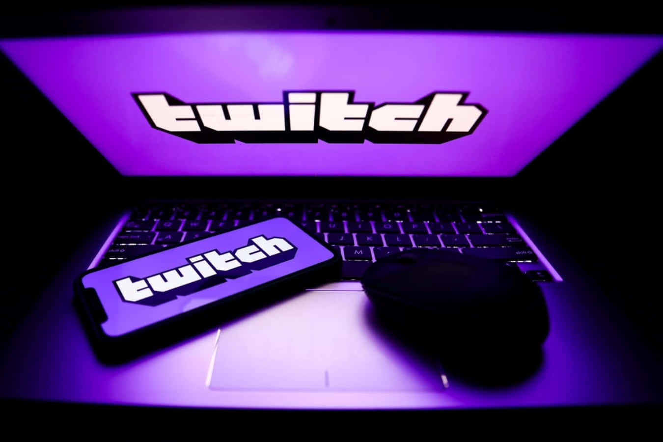Twitch 桌布 壁紙 — 一台筆記型電腦和智慧型手機顯示Twitch標誌，沐浴在充滿活力的紫色光線中，背景為深色。