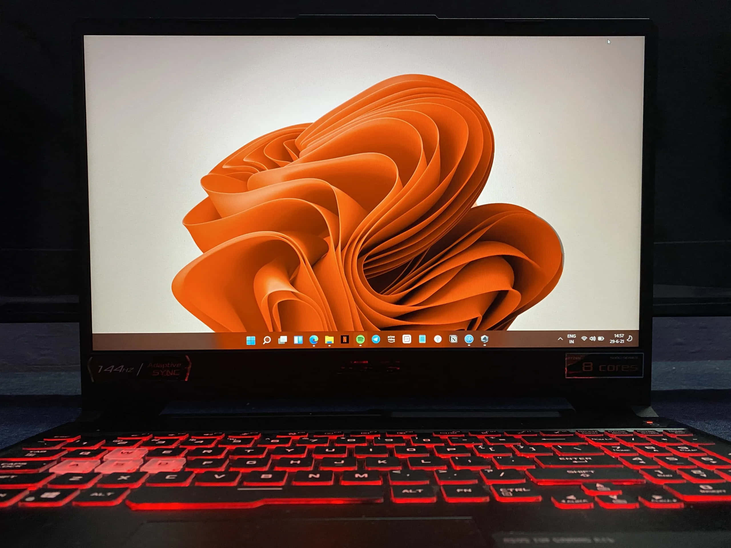 ASUS 桌布 壁紙 — 一台筆記型電腦在純白背景上顯示充滿活力的橙色抽象漩渦桌布，下方有紅色背光鍵盤。