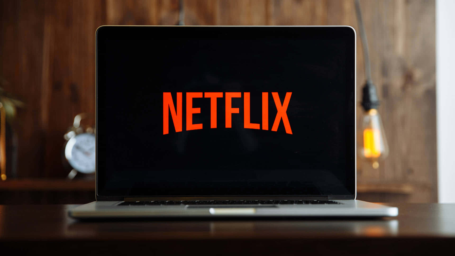 筆電 桌布 壁紙 — 一台筆記型電腦在黑暗螢幕上顯示明亮的橙色Netflix標誌，背景是溫暖的木桌，桌上有一個發光的燈泡和一個時鐘。
