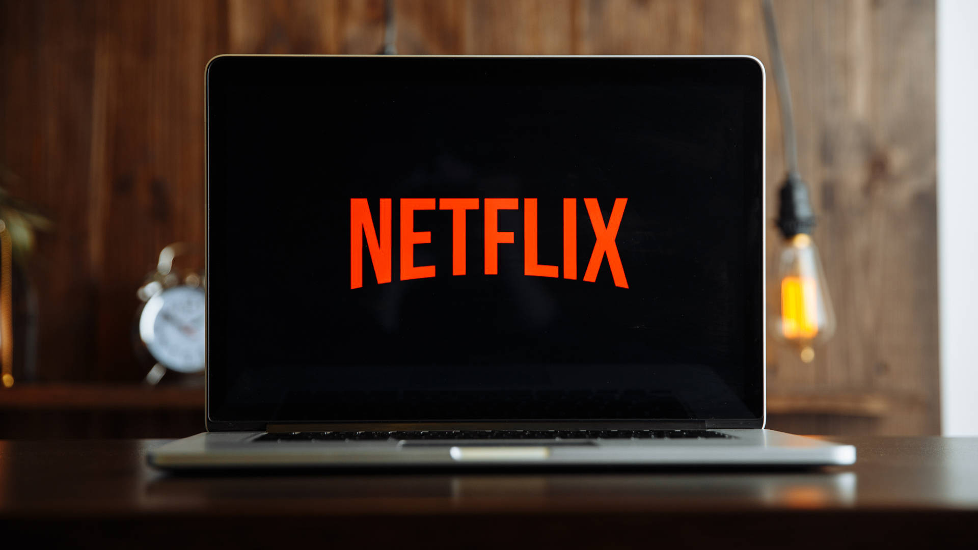 Netflix 桌布 壁紙 — 一台筆記型電腦在黑暗螢幕上顯示明亮的橙色Netflix標誌，放置在木桌上，配有一個復古鬧鐘和發光的愛迪生燈泡，在溫暖的鄉村風格房間中。
