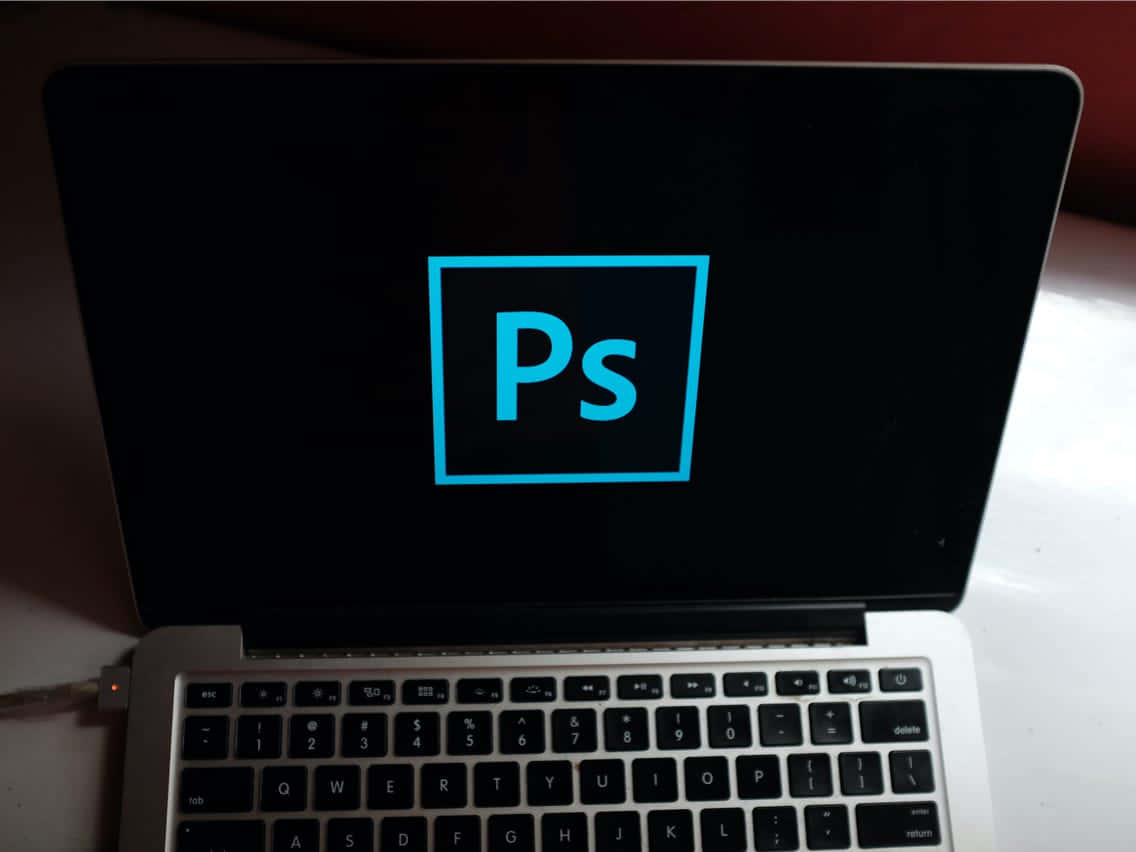 筆電 桌布 壁紙 — 一台筆記型電腦在黑色螢幕上顯示發光的藍色Adobe Photoshop標誌，下方可見鍵盤。